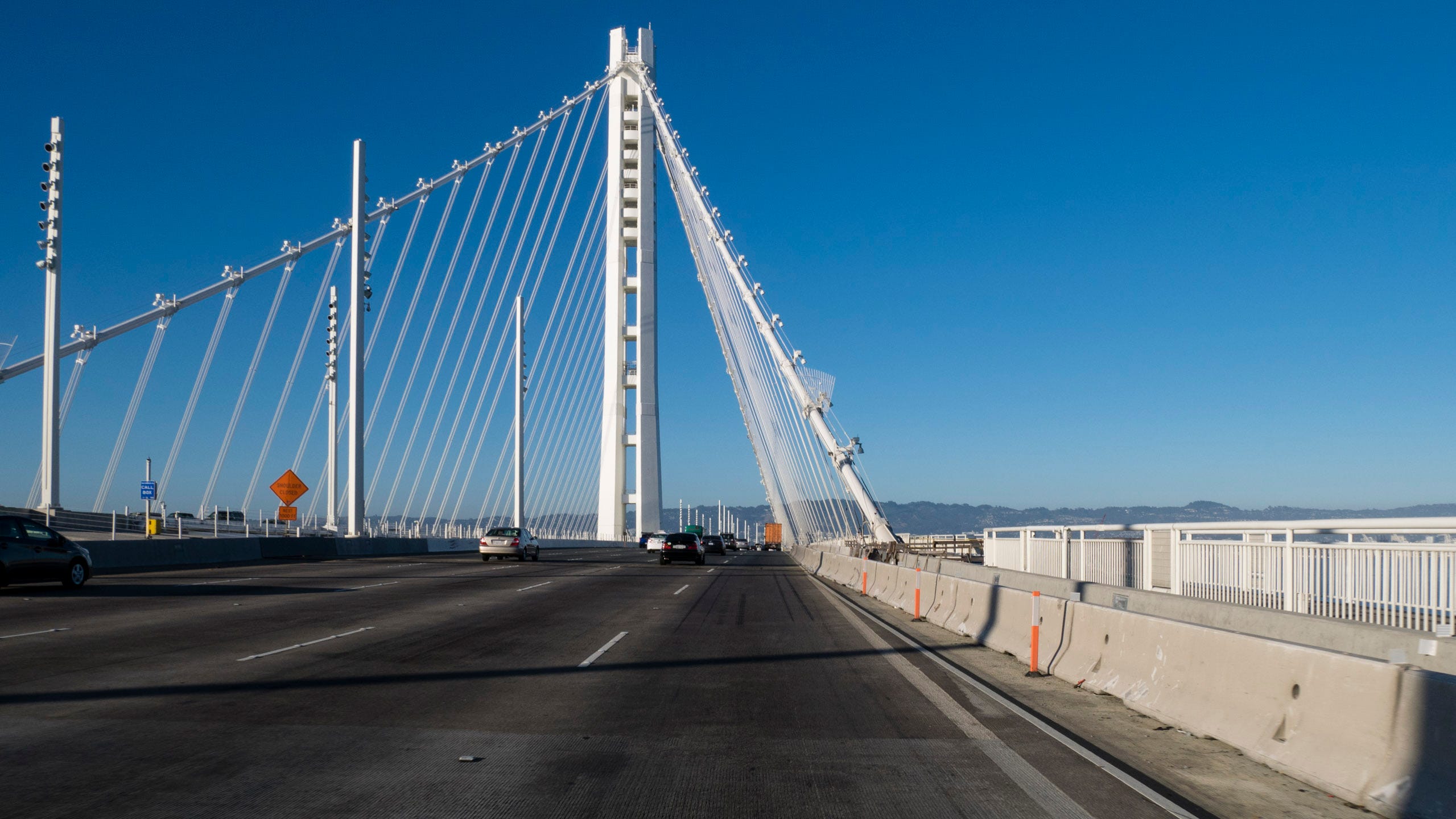 Photo from the right lane of the Bay Bridge. Ahead is a futuristic, gleaming white structural tower from which four thick support cables descend at approximately 45 degree angles to the bridge deck, creating a pyramidal shape of the structural units. Thinner cables extend downward from the four main cables towards the bridge deck.