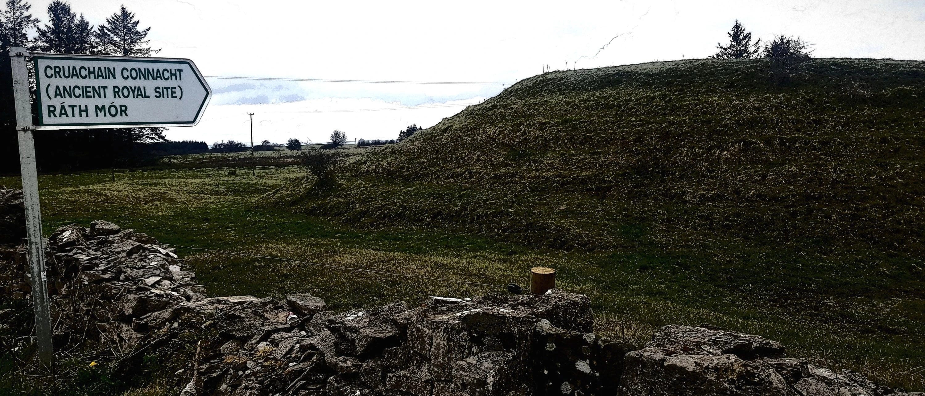 The mound of Rath Mór on the other side of a stone wall with a white road sign stating "Cruachaain Connacht (ancient royal site) Rath Mór"