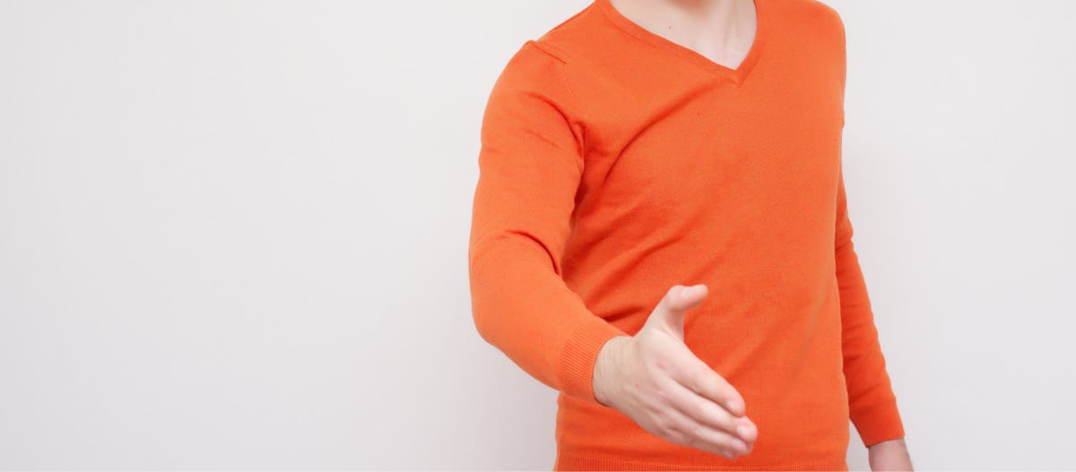 Stock photo wih a grey-white background. The image captures the torso, from about the collar bone down, of a white man in an orange v-neck jumper. His hand is extended, as if offering it to shake. 
