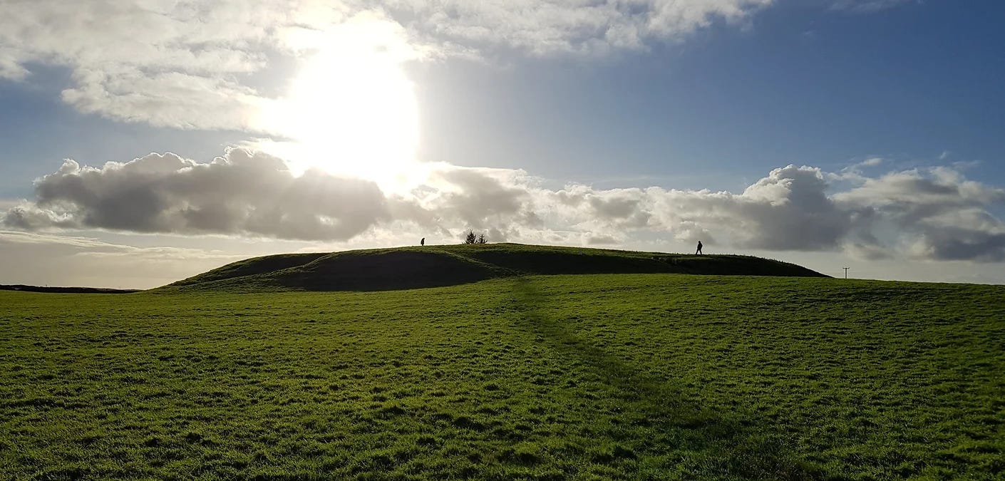 The large flat-topped mound of Cruachan where Queen Medb was reputed to have held court, set in grassy field, with two people walking on the top, against a blue sunlit sky