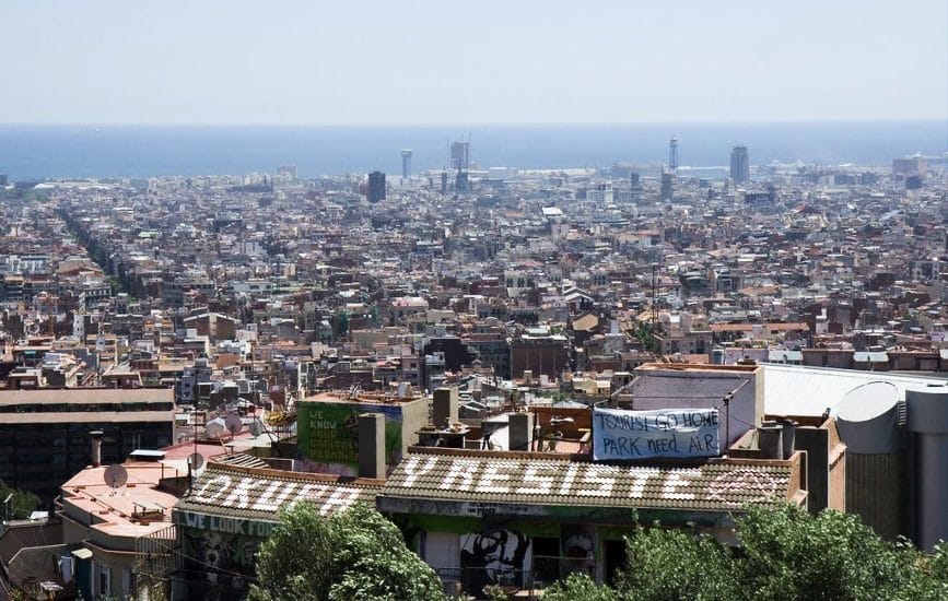 Una panoramica di Barcellona dall'alto e dei messaggi di protesta su alcuni tetti, che dicono Tourist go home e Okupa y resiste