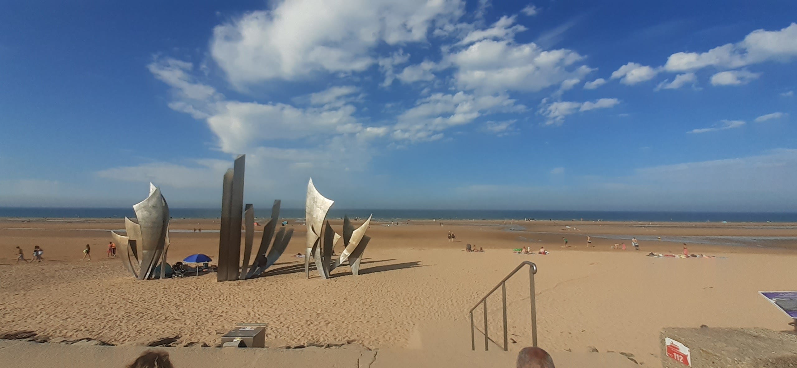 La lunghissima spiaggia di Omaha Beach in un pomeriggio di agosto: il cielo è azzurro e attraversato da poche nuvole, il mare si scorge in lontananza. Sulla spiaggia diverse persone prendono il sole. Sulla sinistra, tre alte sculture astratte in metallo ricordano i caduti del D-Day.