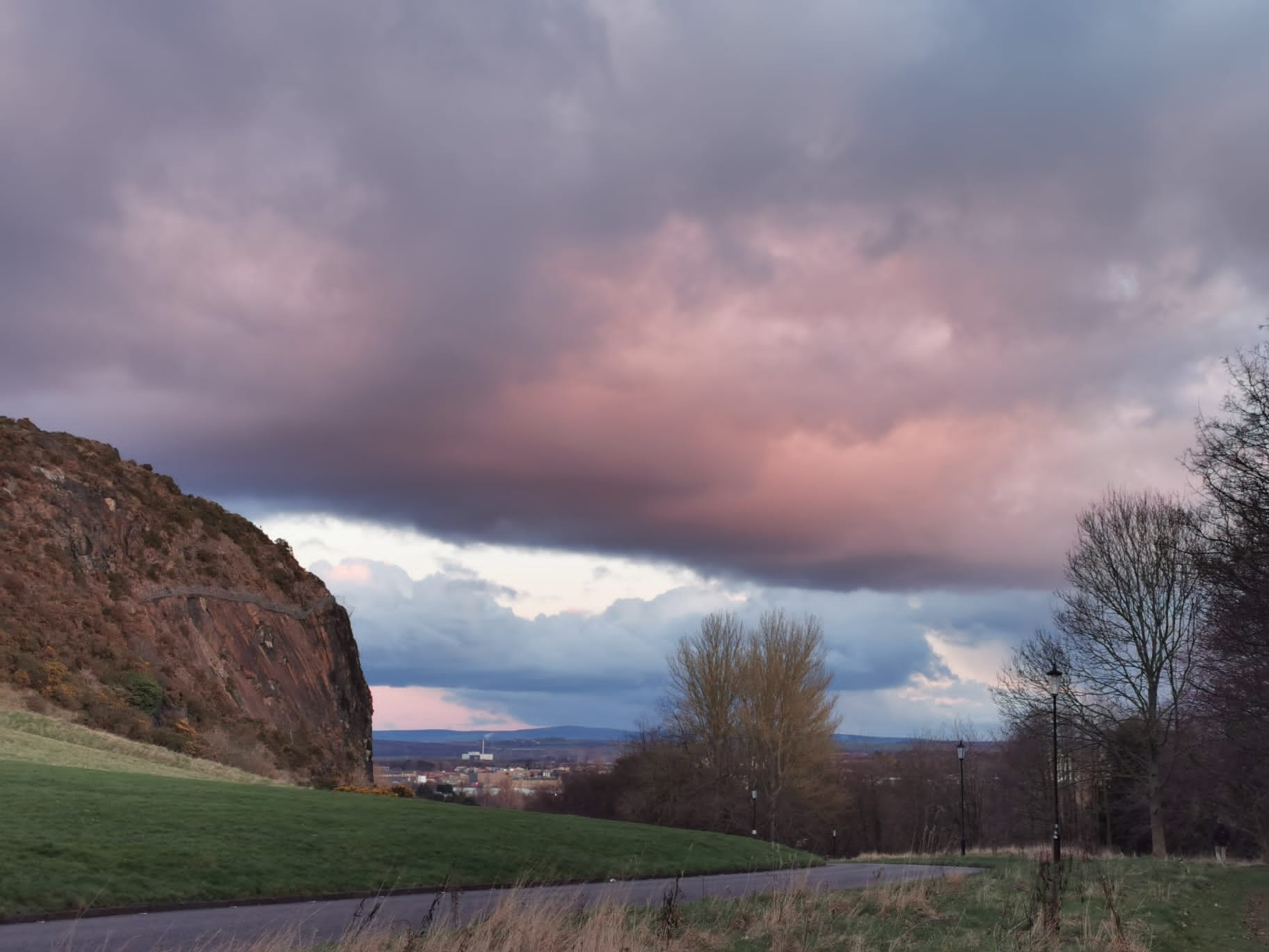 Pink and grey clouds cover the sky, with a view of a hill to the left and road curving down to houses below. There are no leaves on the trees. 