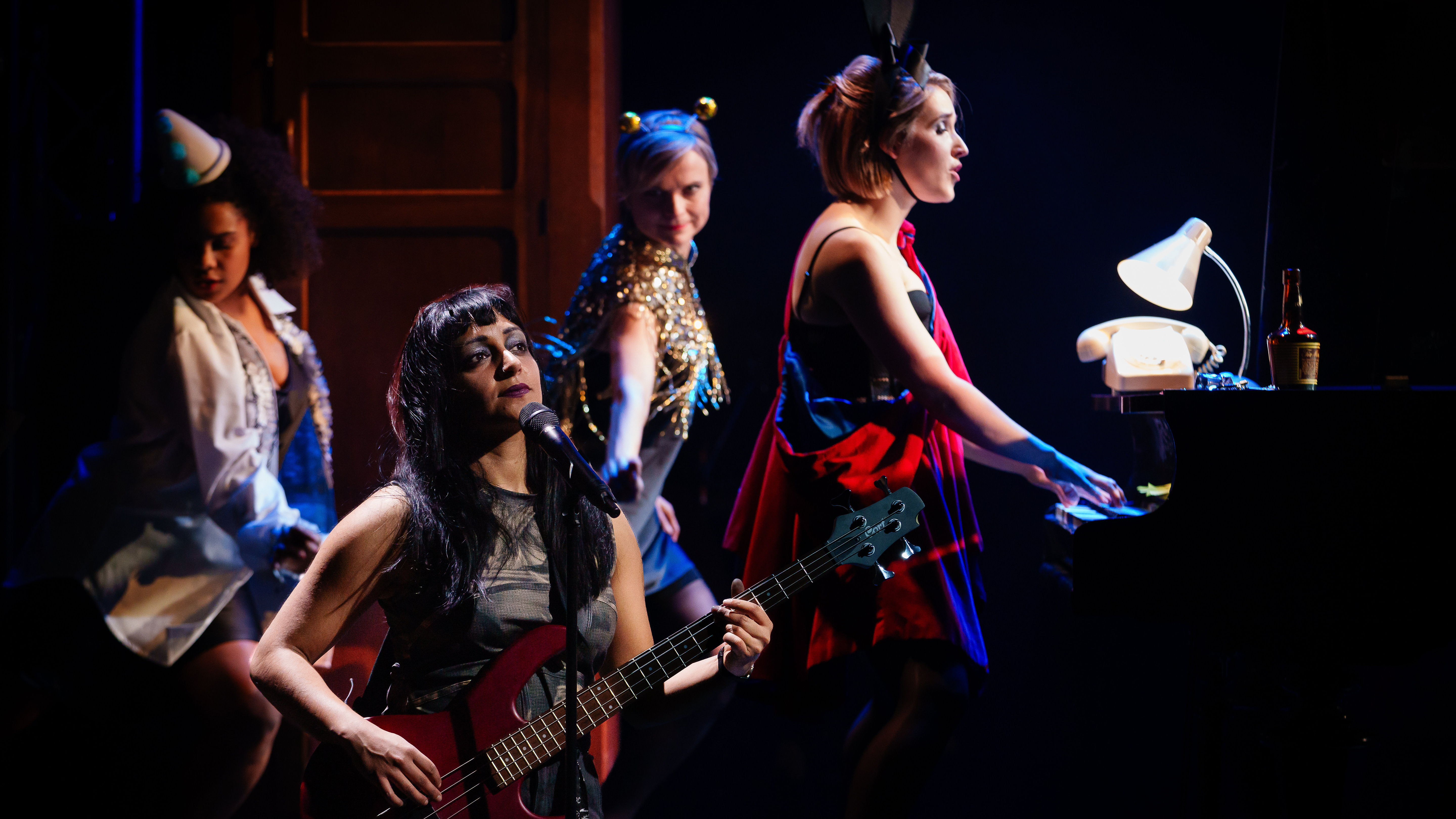 Three women in fancy dress dance behind a Goth woman playing a guitar