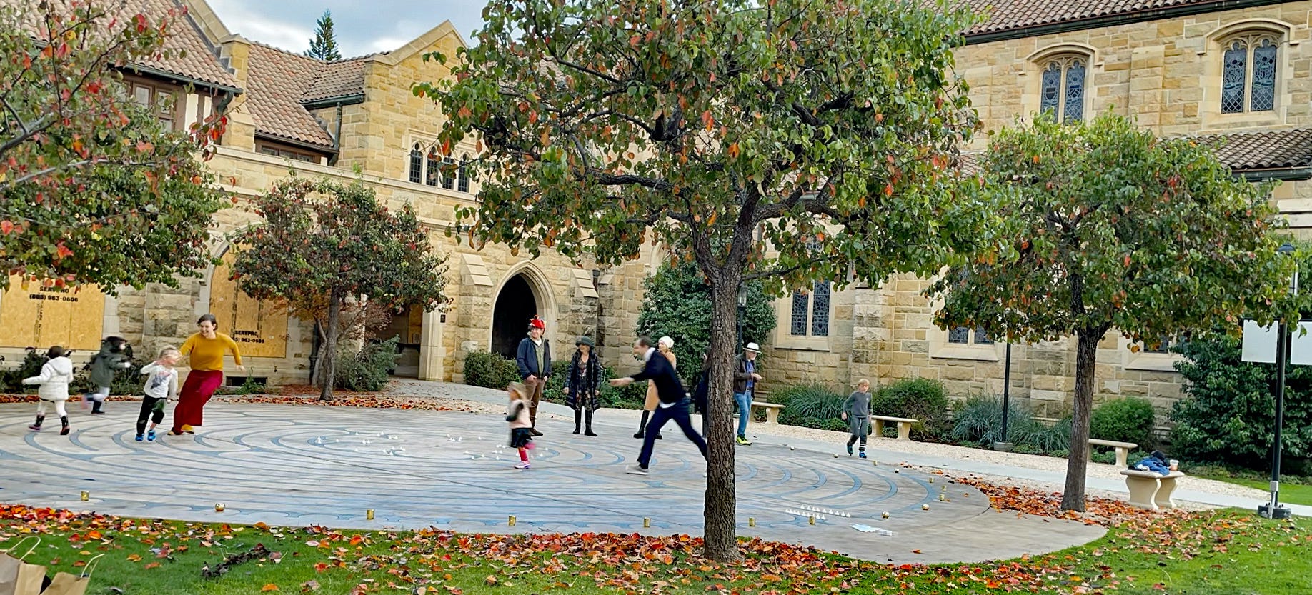 Members of a large family walk around on a stone labyrinth in a church courtyard. Children are running and adults chasing them. 