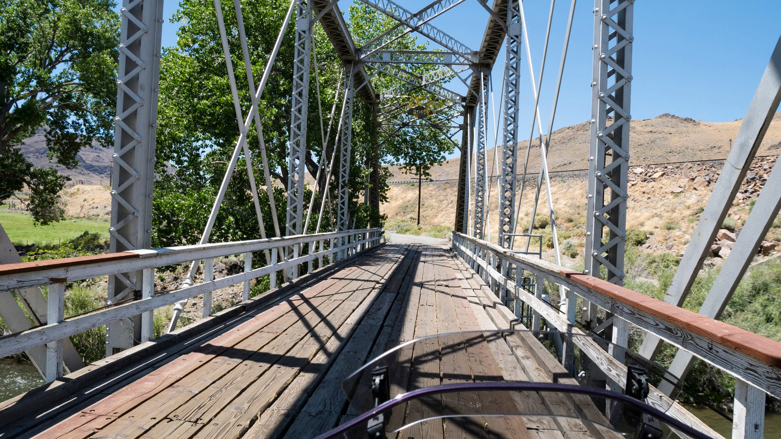Photo taken from the seat of a motorcycle of the bike crossing an old, single-lane wooden-decked truss bridge. The vertical columns and diagonal braces of the bridge fill the frame and cast strong shadows onto the bridge deck. To the left, leafy, green trees grow alongside the river that the bridge is crossing. Beyond, is a green, grassy field and then dry, arid desert. To the right, elevated above the roadway, the level bed and rail of a railroad track can be seen. Beyond that, a naked, rounded hill. Straight ahead is the end of the bridge. The road bends to the left. A single telephone pole can be seen on the side of the road.