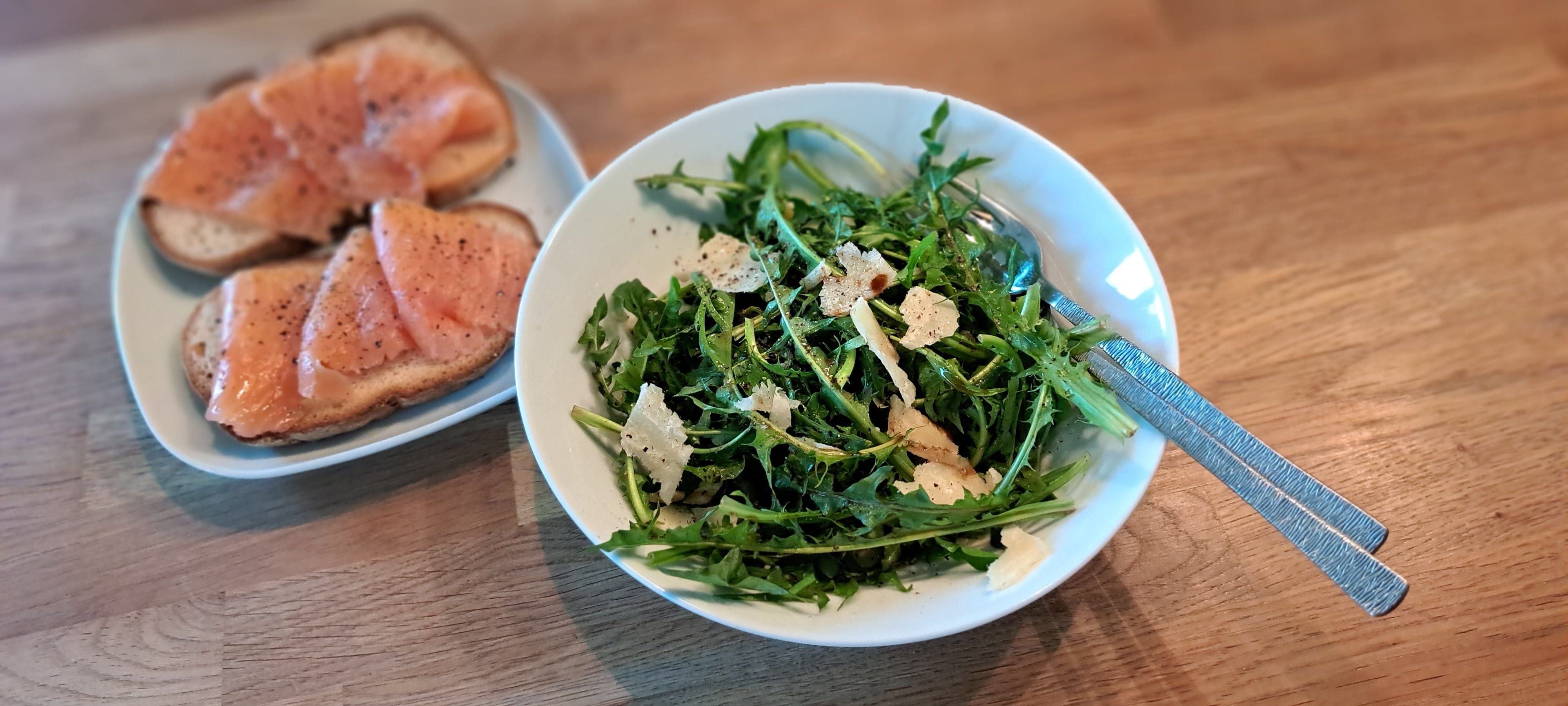 A white bowl containing a salad of jagged dandelion leaves sprinkled with salt and pepper and shavings of parmesan. Beside is a platw with toast and smoked salmon.