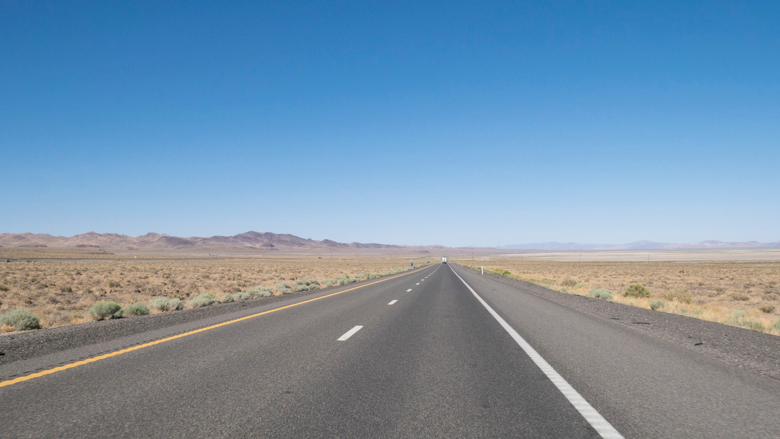 Photo of riding on the highway across the dry and barren Forty Mile Desert in Nevada. The sky is crystal blue. The highway heads arrow straight toward a vanishing point on the horizon, where barren brown mountains lie. Alongside the road is sand and sage.