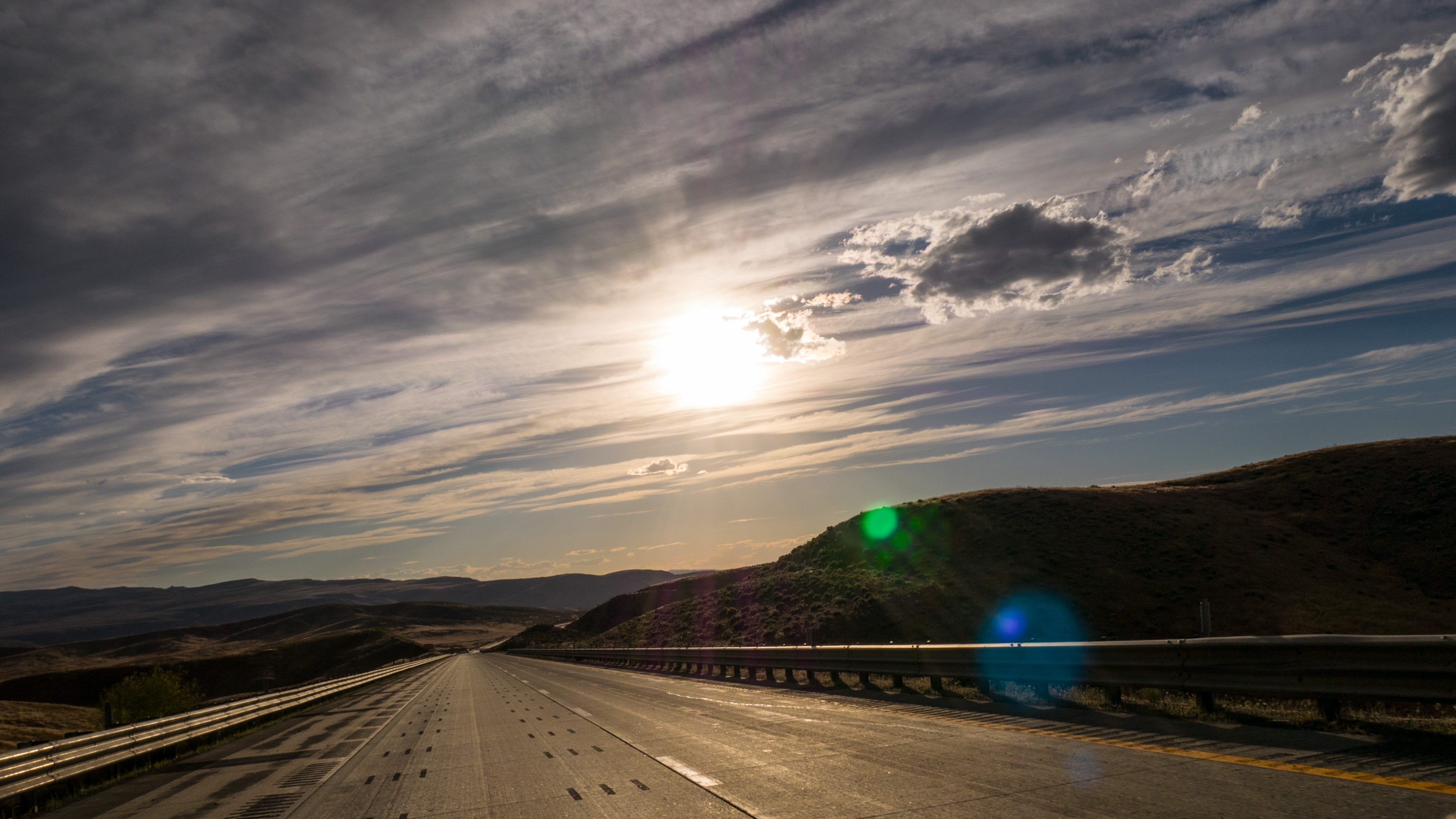 Photograph looking westward along Interstate 80 in Nevada. The road inclines downward to a valley before turning slightly and climbing towards a distant set of rugged mountains. It is late afternoon and the light is low, a strong sun in a blue sky streaked with clouds. The landscape is dark, in silhouette and the sunlight reflects off the asphalt of the highway. The highway guardrail glistens in the strong light.
