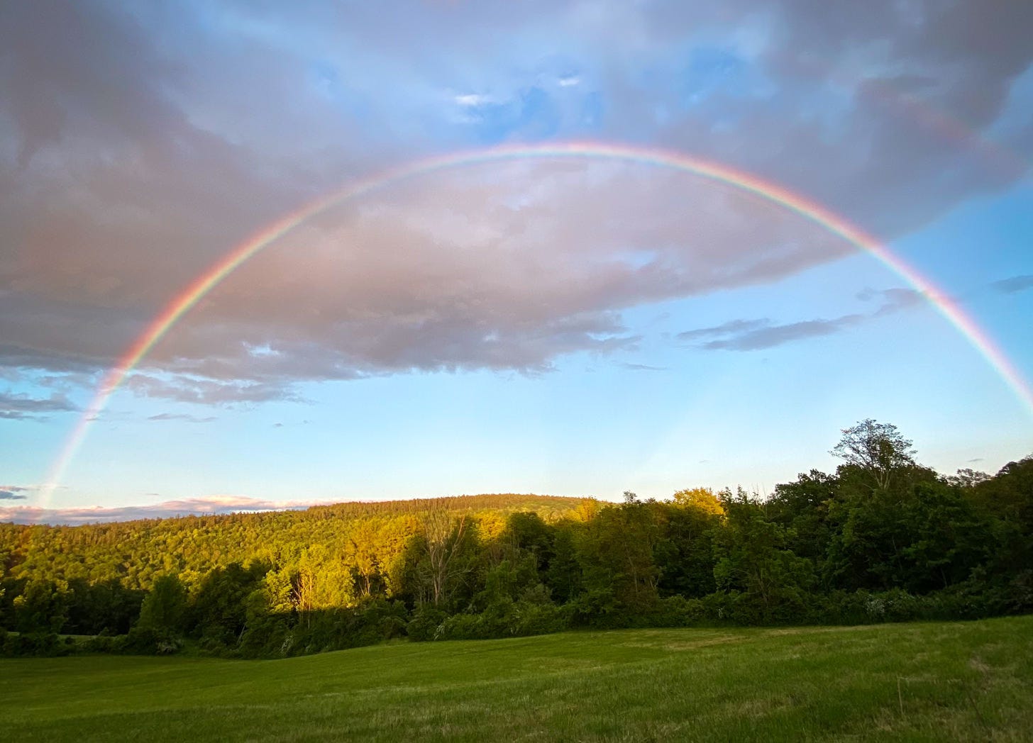 rainbow against blue sky with gray clouds above grassy meadow and wooded hills