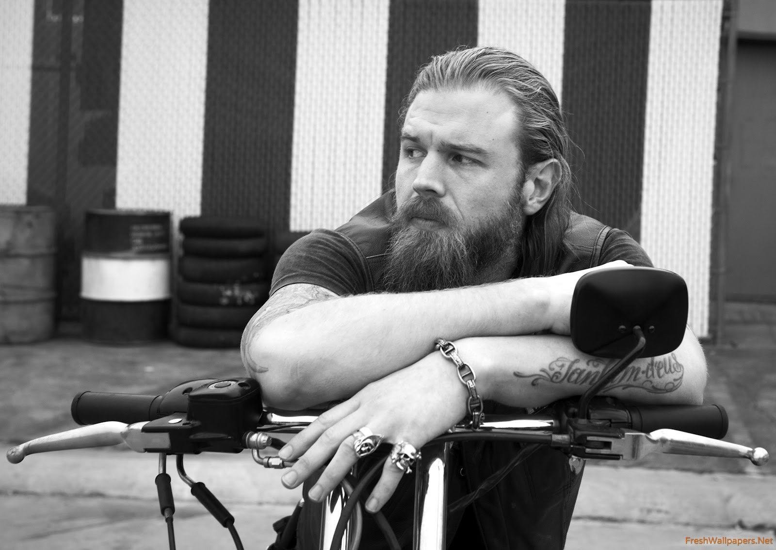 Homem branco, cabeludo e barbudo, de cara séria e anéis de caveira nos dedos, tatuado, sentado numa motocicleta, vestido de preto com colete de couro.