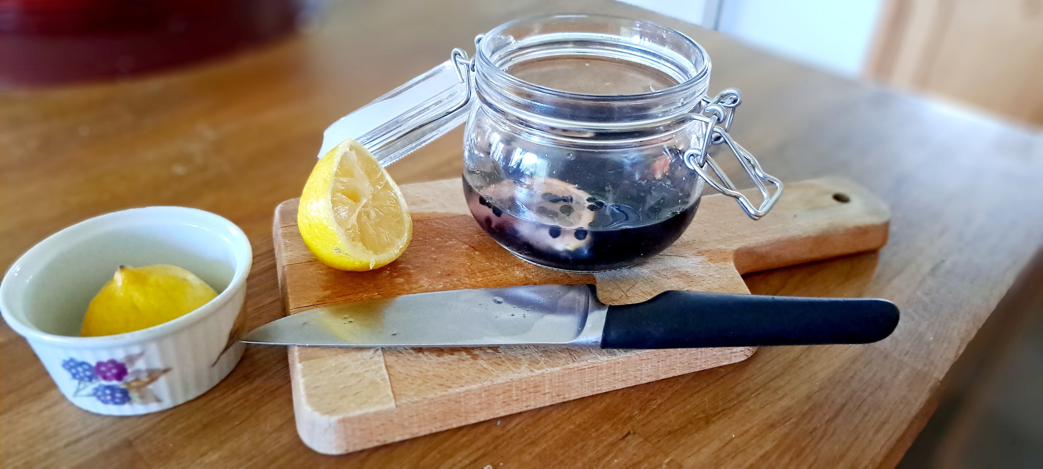 A small jar with top open resting on a wooden chopping board. In front is a knife with a black handle, to the left half a lemon, with the other half in a white ramekin. The jar is half filled with gin, elderberries and a slice of lemon.