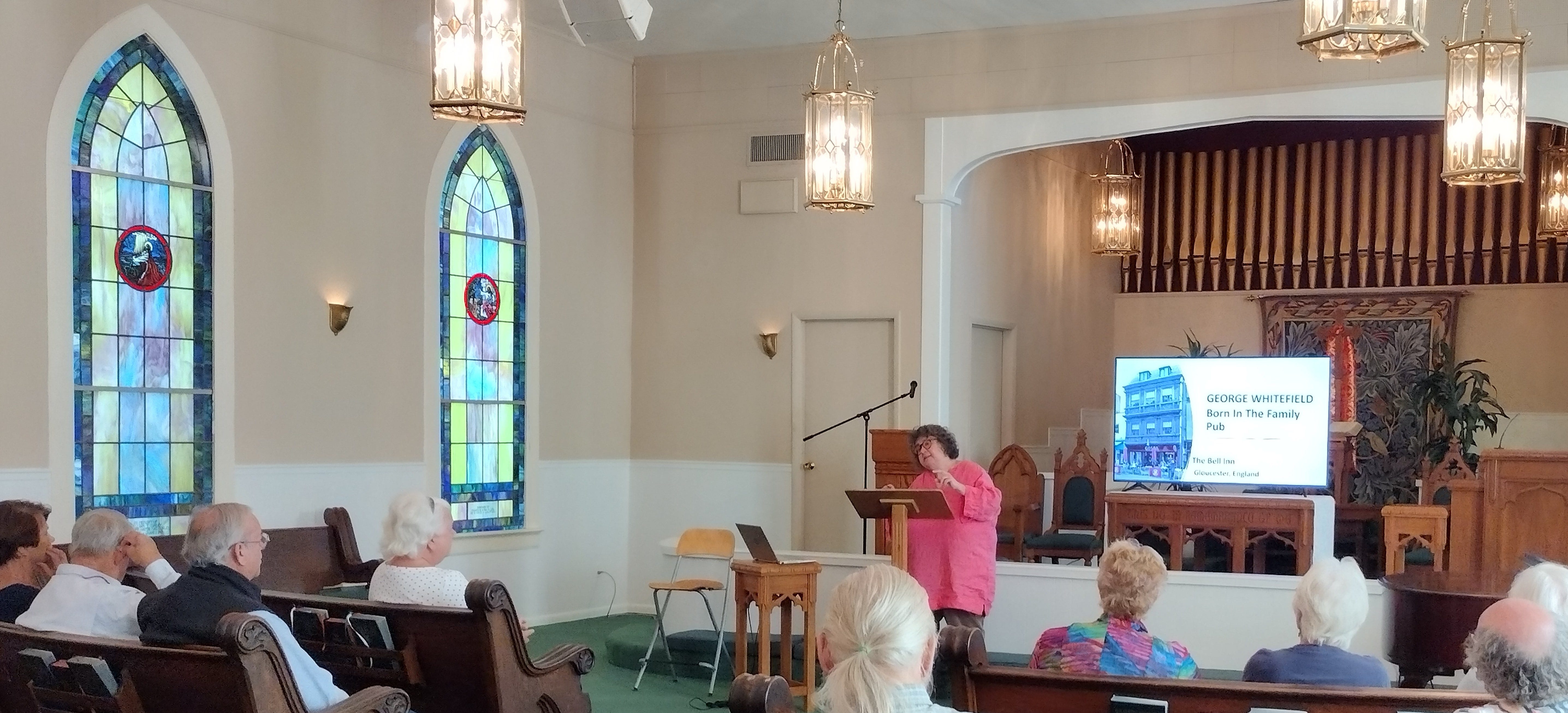 Annette at podium in church with stained glass and pews talking to audience
