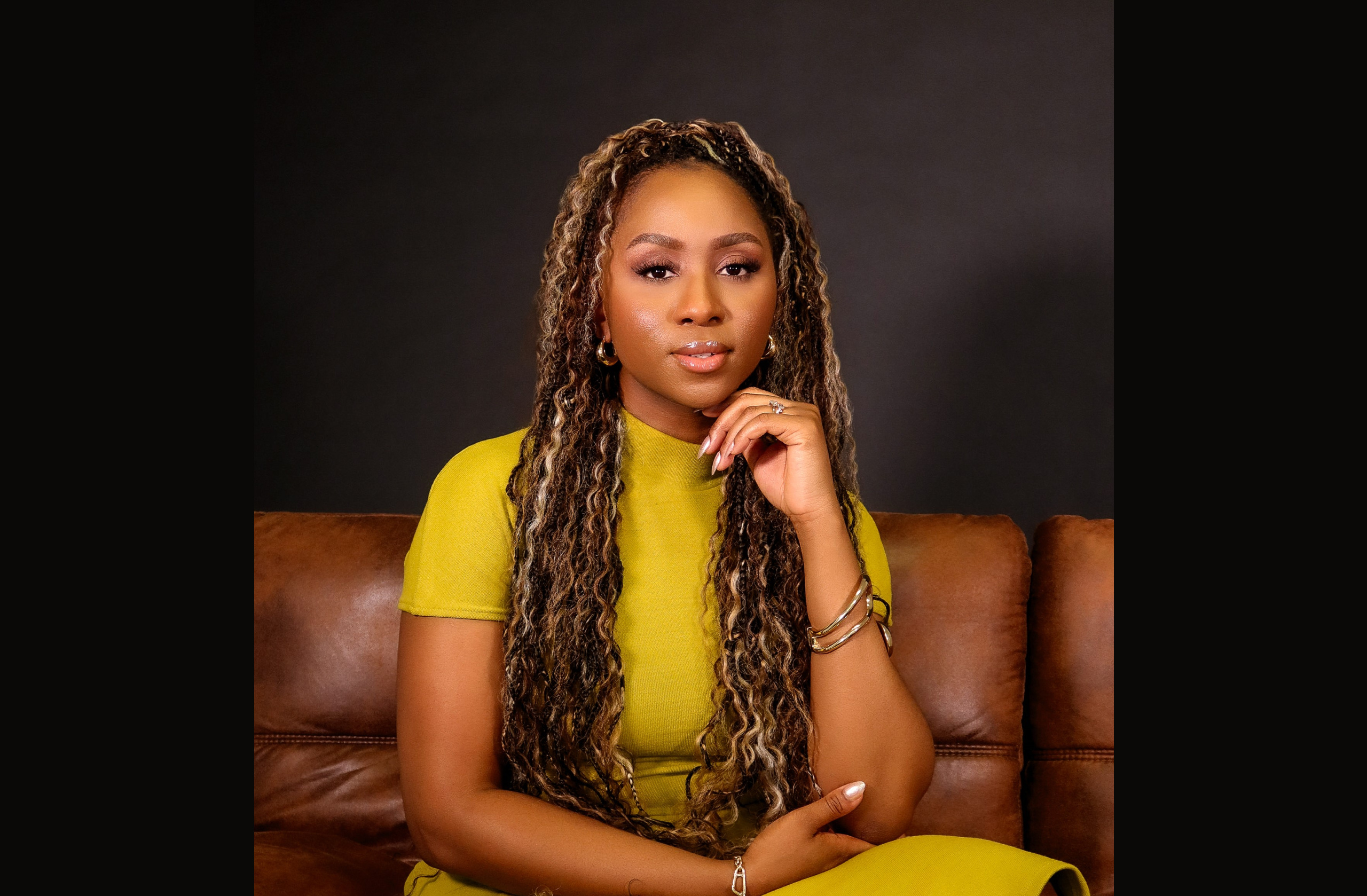 Hope Idaewor sits facing the camera in a yellow dress posing for a portrait on a brown couch.