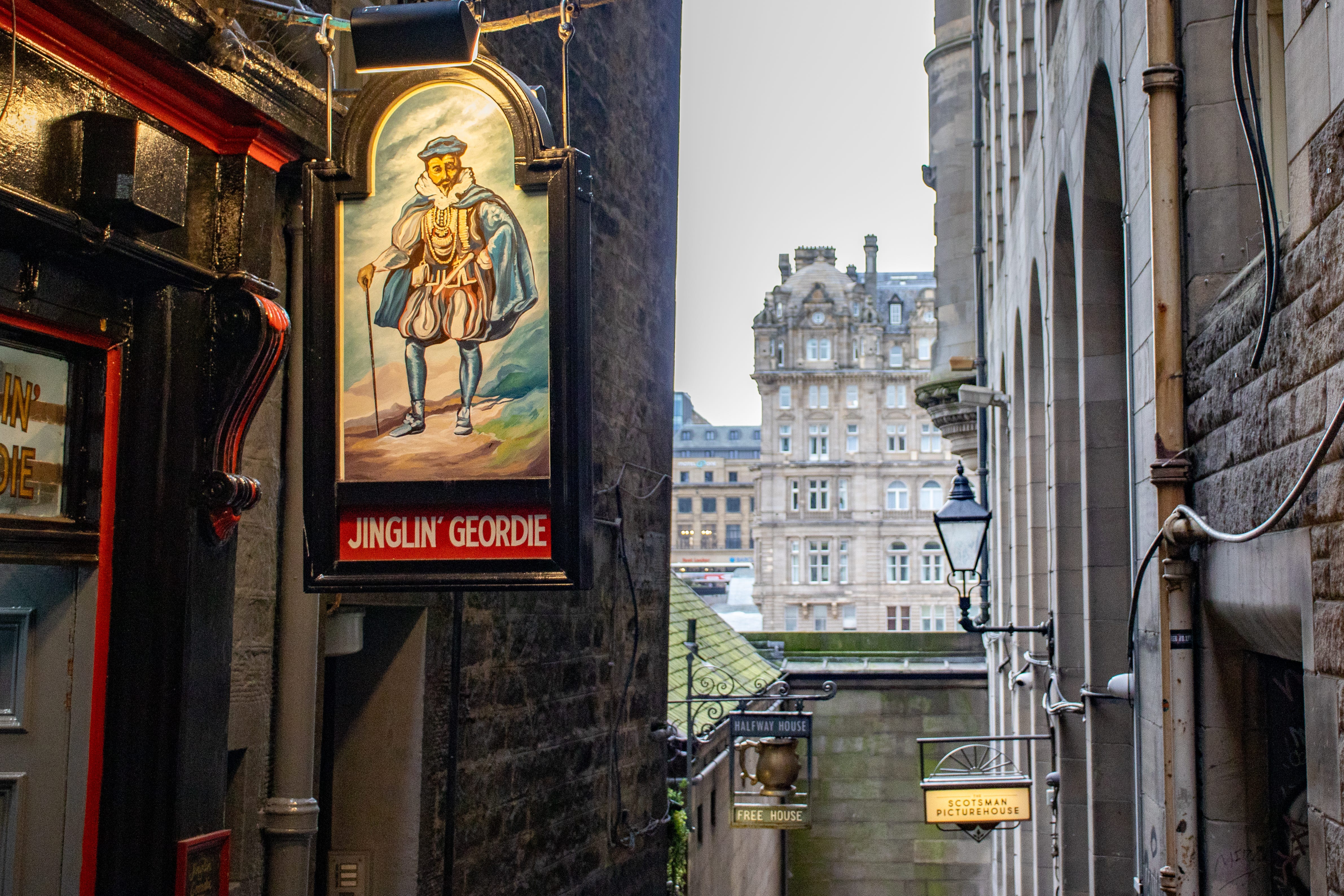 Pub sign depicting a painting of a man in elaborate blue 17th century clothes and a walking stick. In the background of the photo is the ornate Balmoral Hotel building.