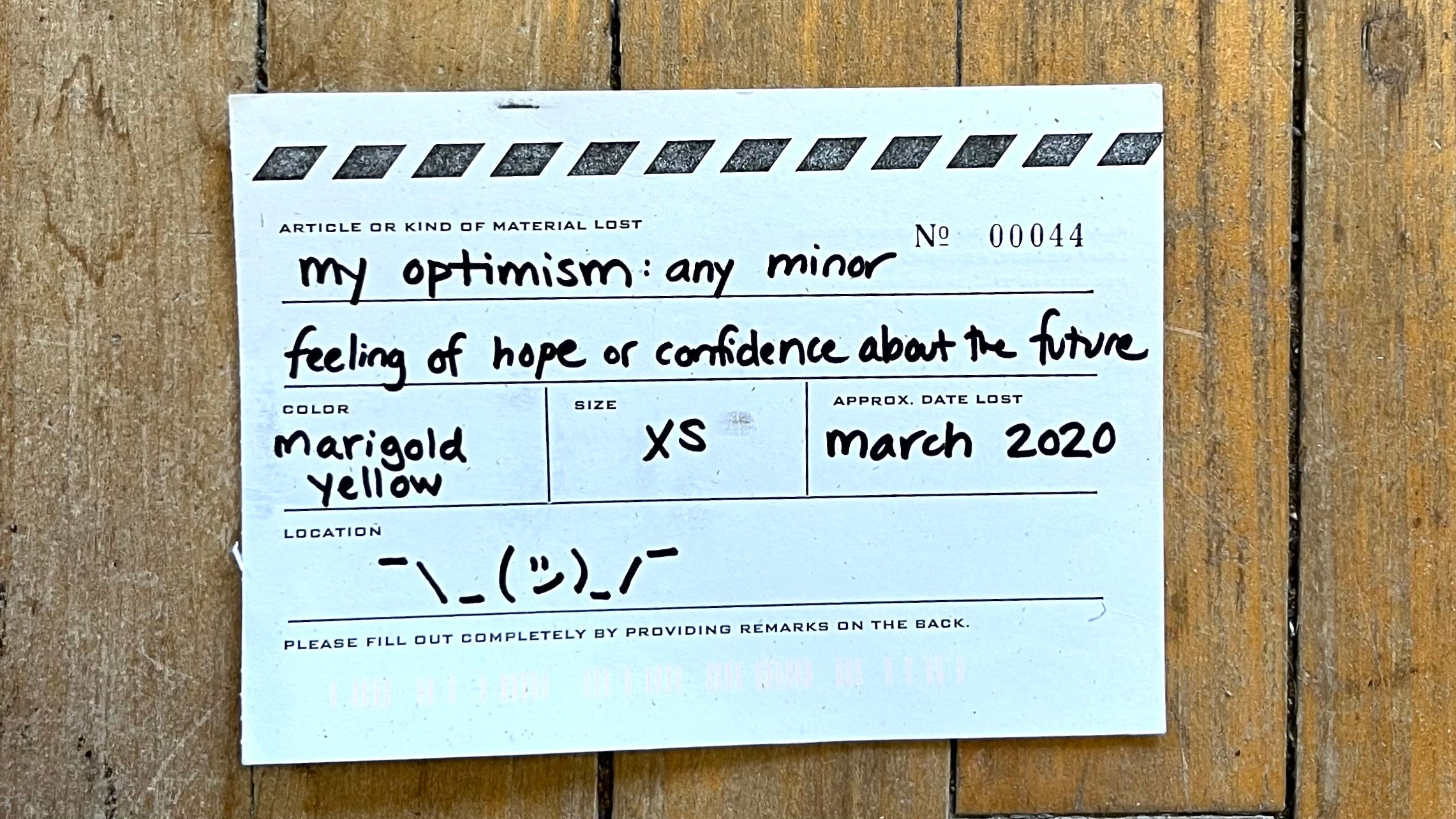 The front of a postcard designed to look like a lost article tag. Under article or kind of material lost it says in handwriting "my optimism: any minor feeling of hope or confidence about the future". Under color it reads "marigold yellow" and under size it says "xs". Approx. date lost reads "march 2020" and under location is the emoticon ¯\_(ツ)_/¯