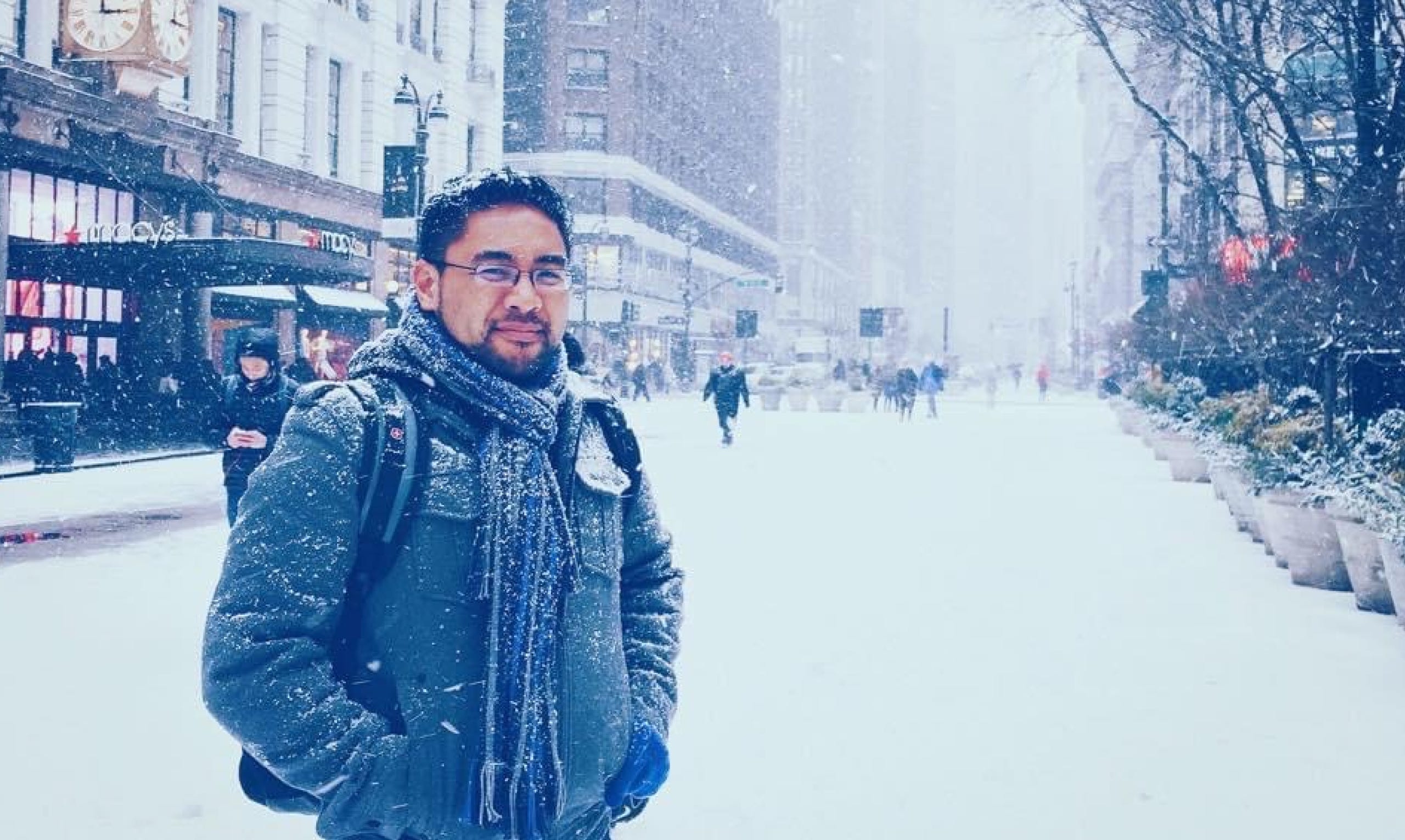 Jay Demetillo in a gray scarf and jacket standing in the street in New York City while it's snowing.