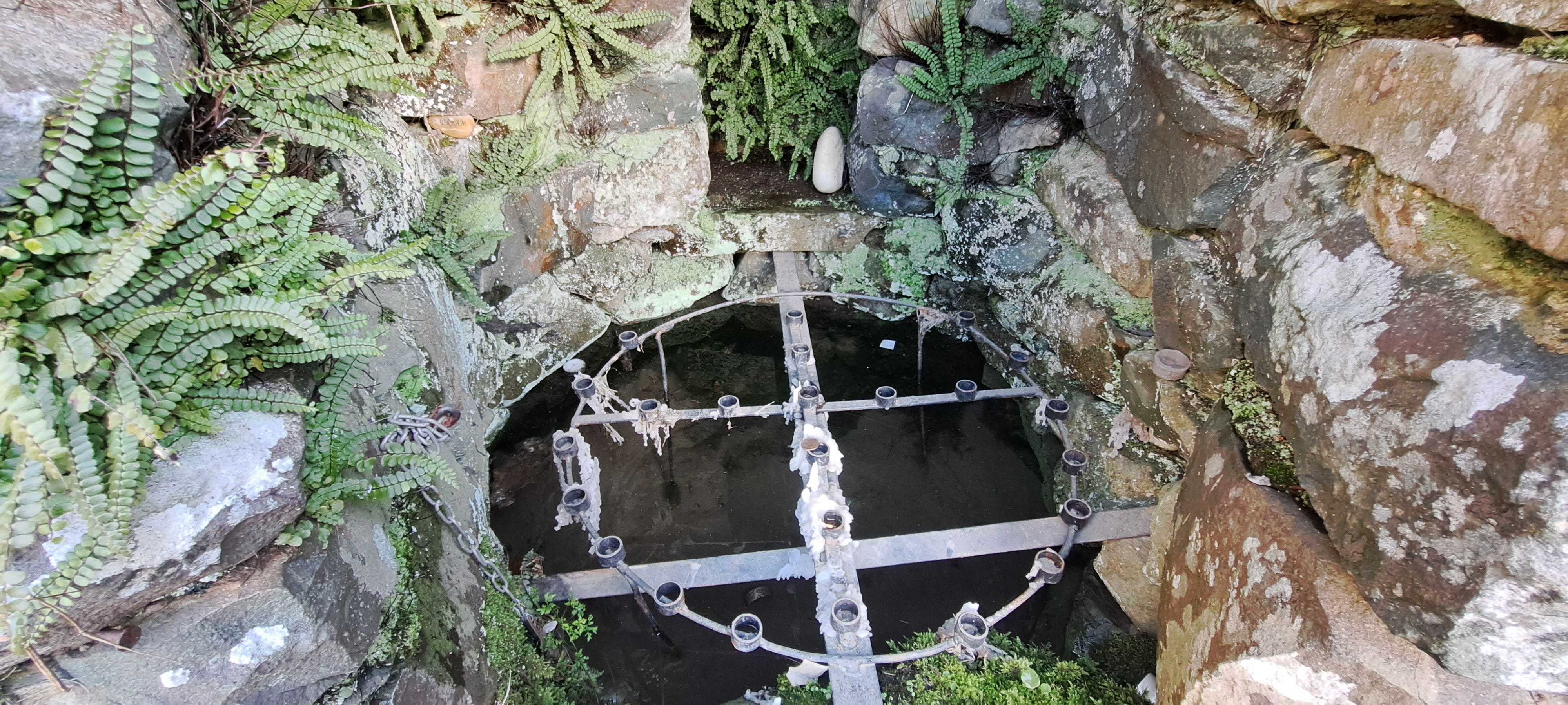 Inside Brigid's well at Raffony: still dark pool of water is enclosed within a rough stone walled enclosure, an iron sconce is suspended directly above the water for holding many candles, dripping wax has set, some tealights are floating in the water, the walls have ferns growing out of them.
