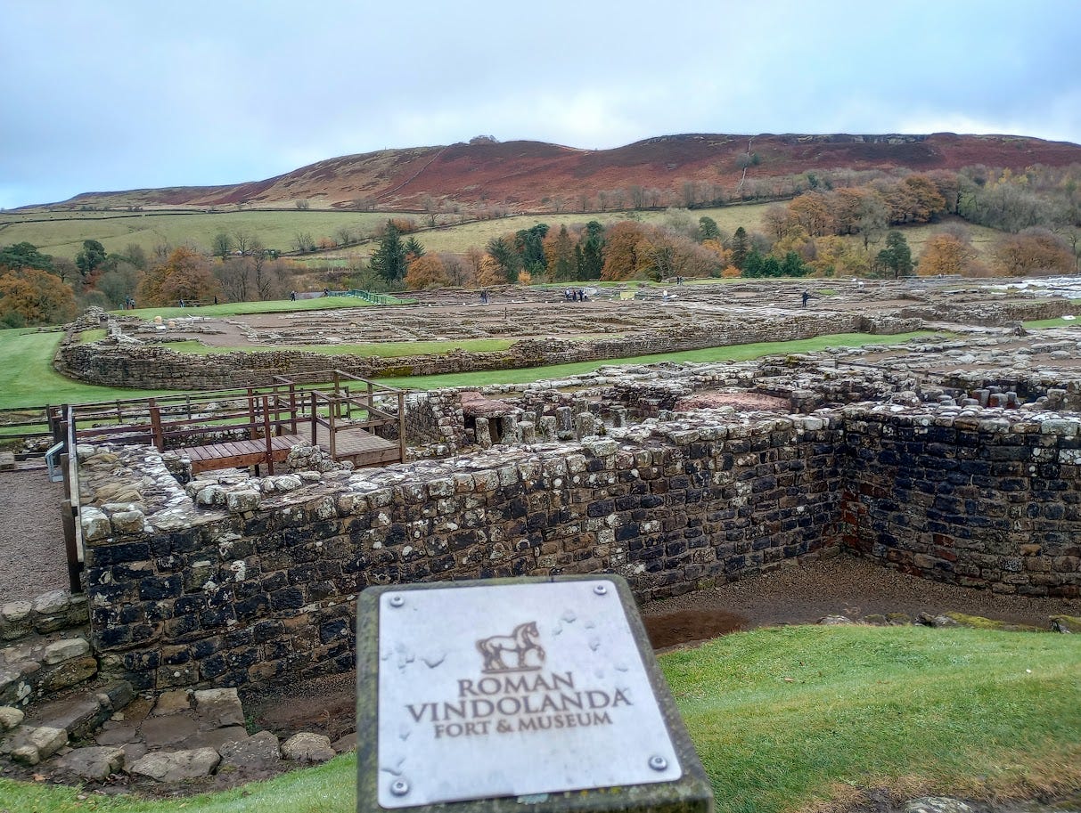 Stone foundations of fort and part of town, with Vindolanda sign, brown and green hills in background