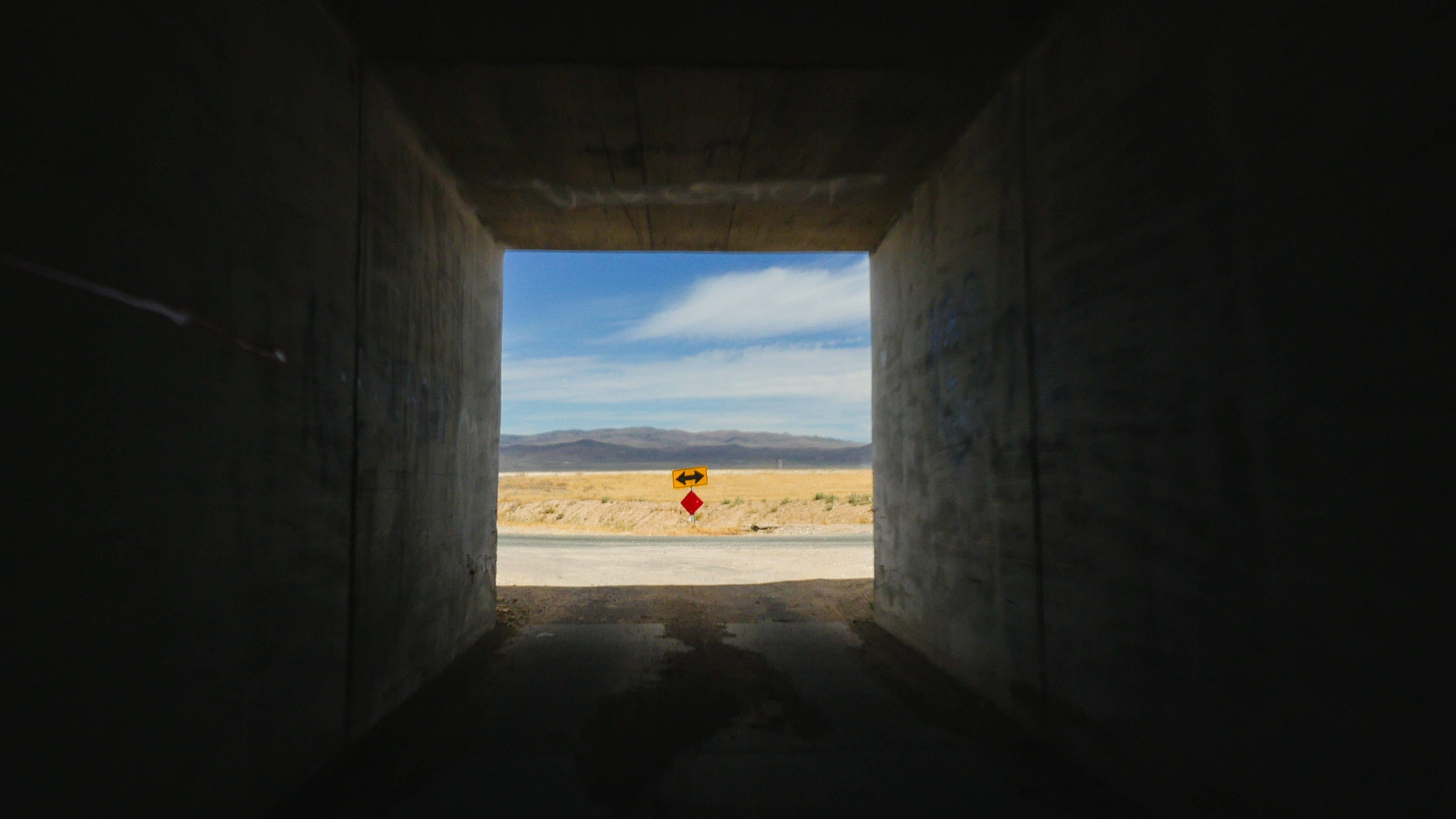 Photo from inside a small, dark square tunnel, looking out the end of the tunnel towards the desert. A road goes left-right at the end of the tunnel. There is a yellow road sign with arrows pointing left and right. Beyond, the short, yellow wild grasses of the Nevada desert, and naked mountains in the distance. Above, the sky is blue and streaked with clouds.