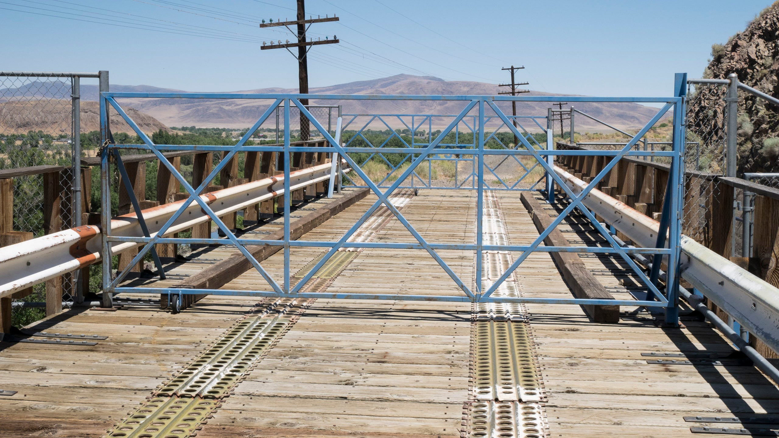 Photo of a wooden-decked bridge with locked gates blocking access. It's a short bridge, with metal tracks where a car or truck's tires are supposed to travel, and white painted guardrails protecting the rusted metal structure of the bridge. About 10 feet from the camera, a metal gate comprised of pieces of blue square tubing laid out in a criss-cross pattern blocks access to the bridge. The gate is hinged on the right; there is a metal chain and lock on the left preventing the gate from swinging open. About 30 feet away from the first gate, a second, identical gate is blocking access. Beyond is low, green scrub brush, a dirt road, and distant, rocky mountains.