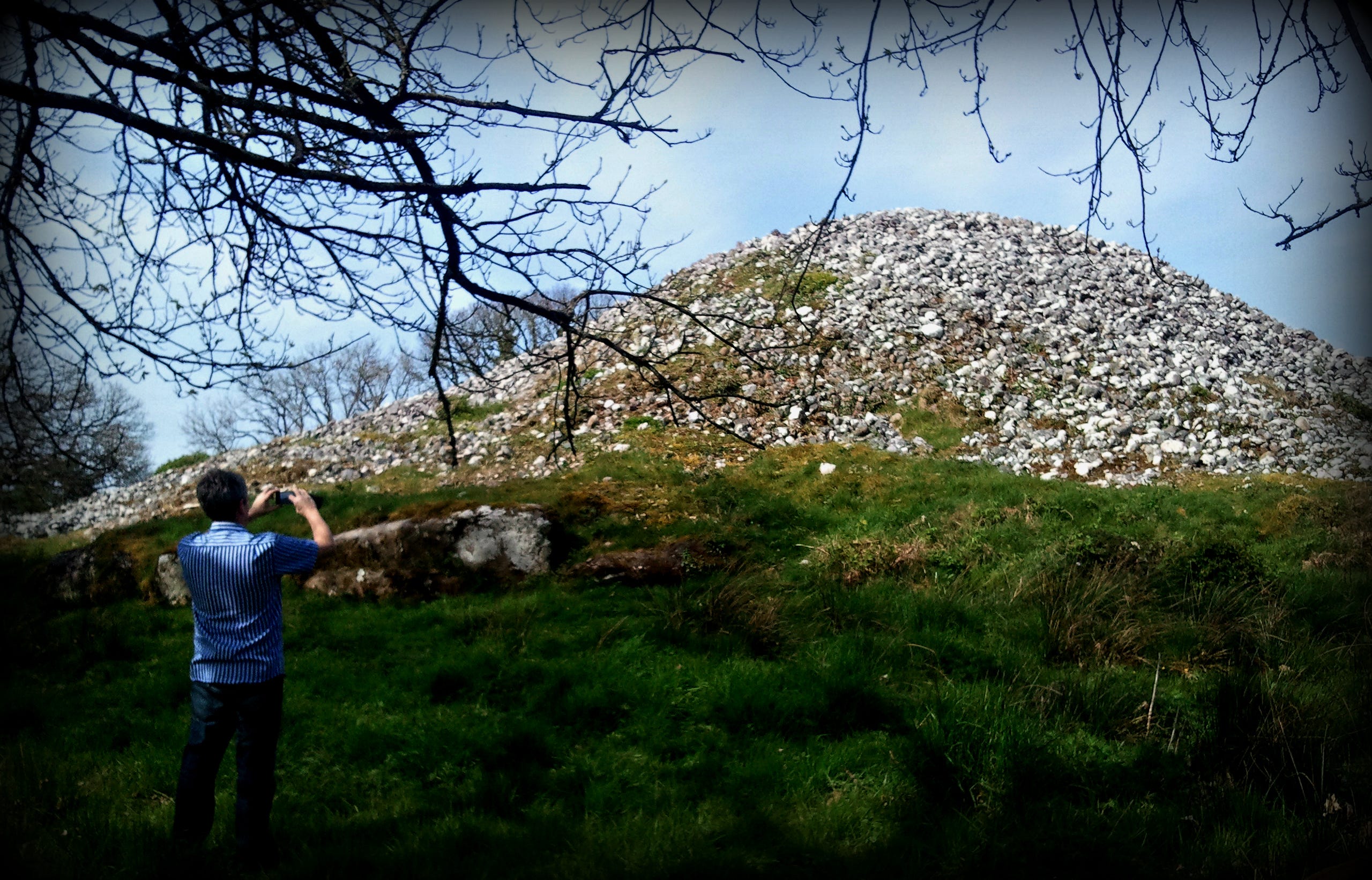 Large cairn covered in small white stones like scree, some large kerb stones visible at the bottom, set against a blue sky, with man dressed in jeans in the foreground beneath a bare-branched tree, taking a photo of the cairn.