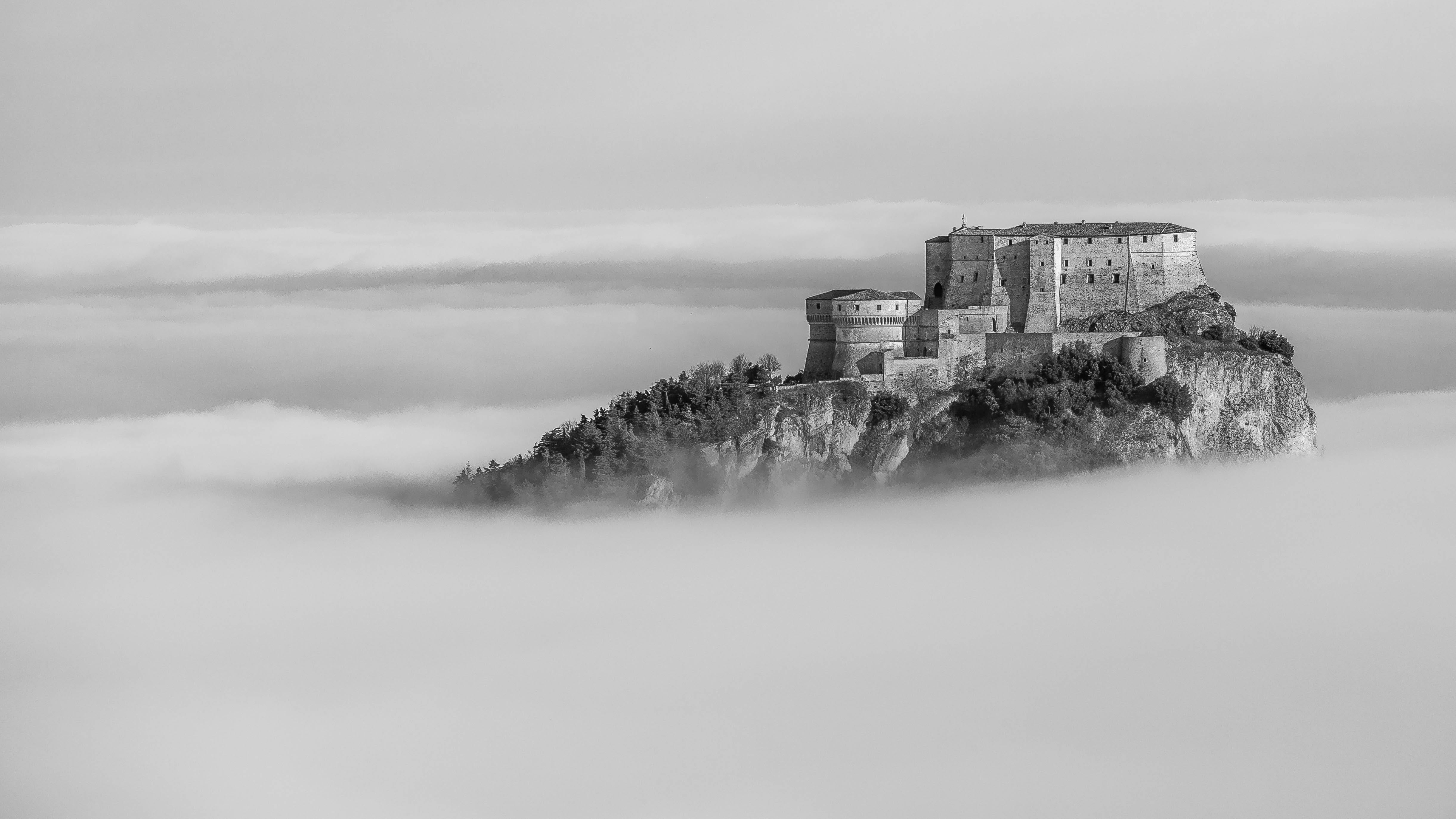 A black-and-white photograph of the fortress of San Leo on a mountaintop above the cloudline.