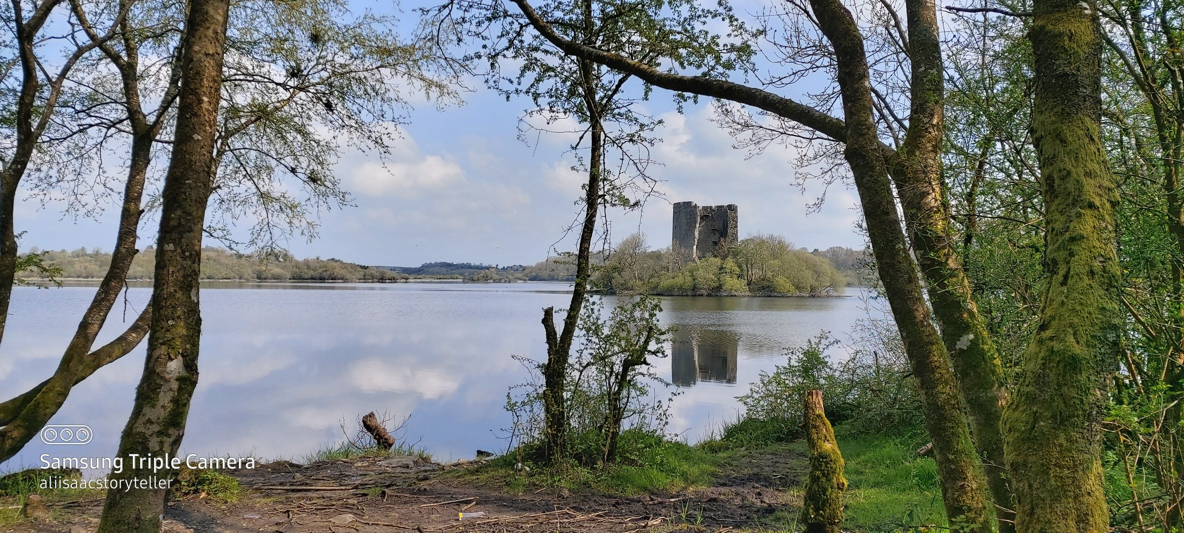 A stine tower-house style castle surrounded by bushes and trees at its base is glimpsed through the trees and branches of Killykeen forest, the sky is pale blue and puffs of cotton wool clouds glide through it, reflected in a mirror-like lake surface. The trees and bushes are greened with new spring growth.