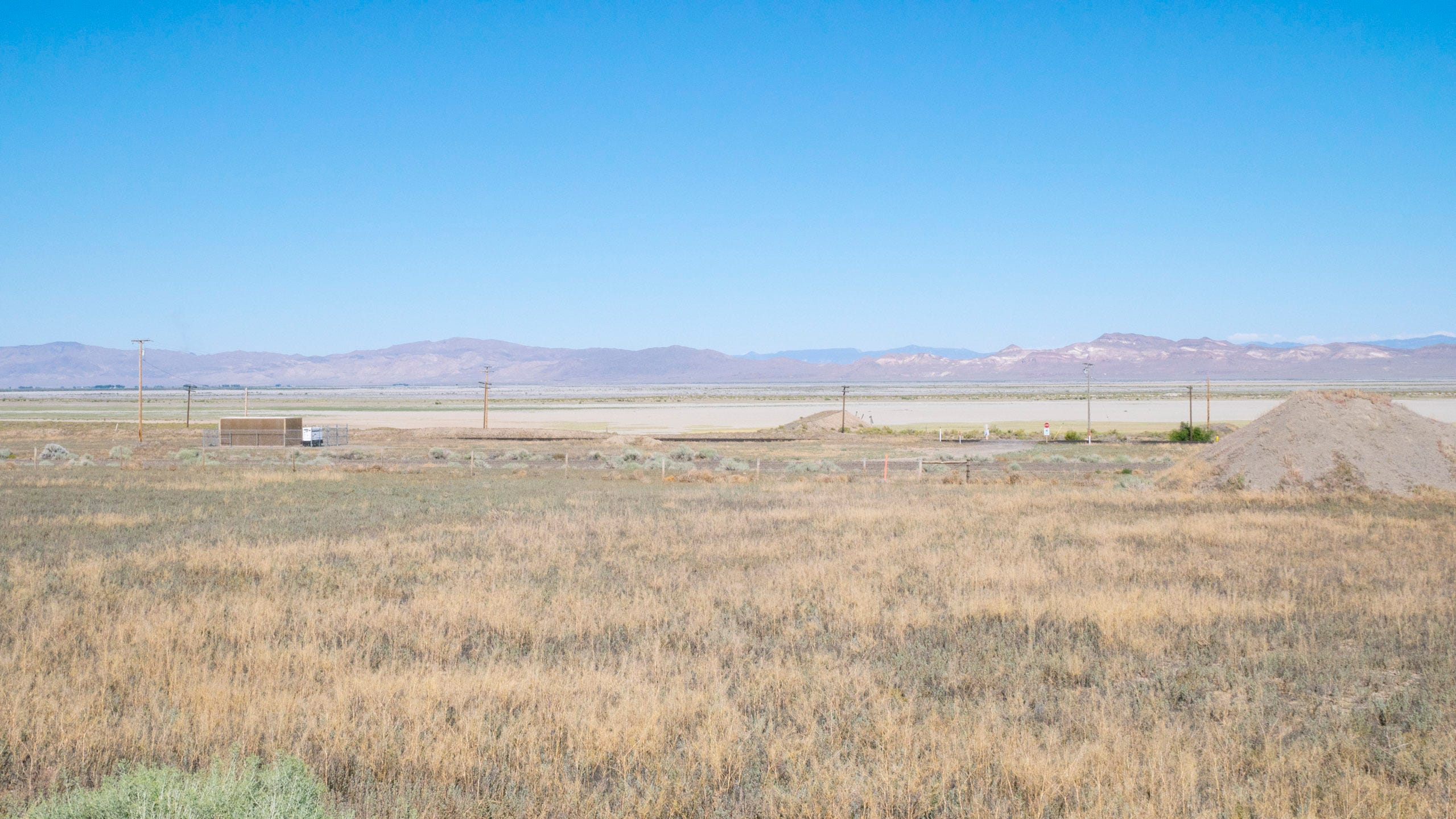 Photograph of the Nevada desert. In the foreground are short, dry grasses. Beyond that, the rails of the railroad can be seen, with telephone poles running alongside and a short, squat utilitarian building (akin to a shipping container) nearby. Beyond, the white sands of the desert. And beyond that in the hazy distance, rugged, barren mountains.