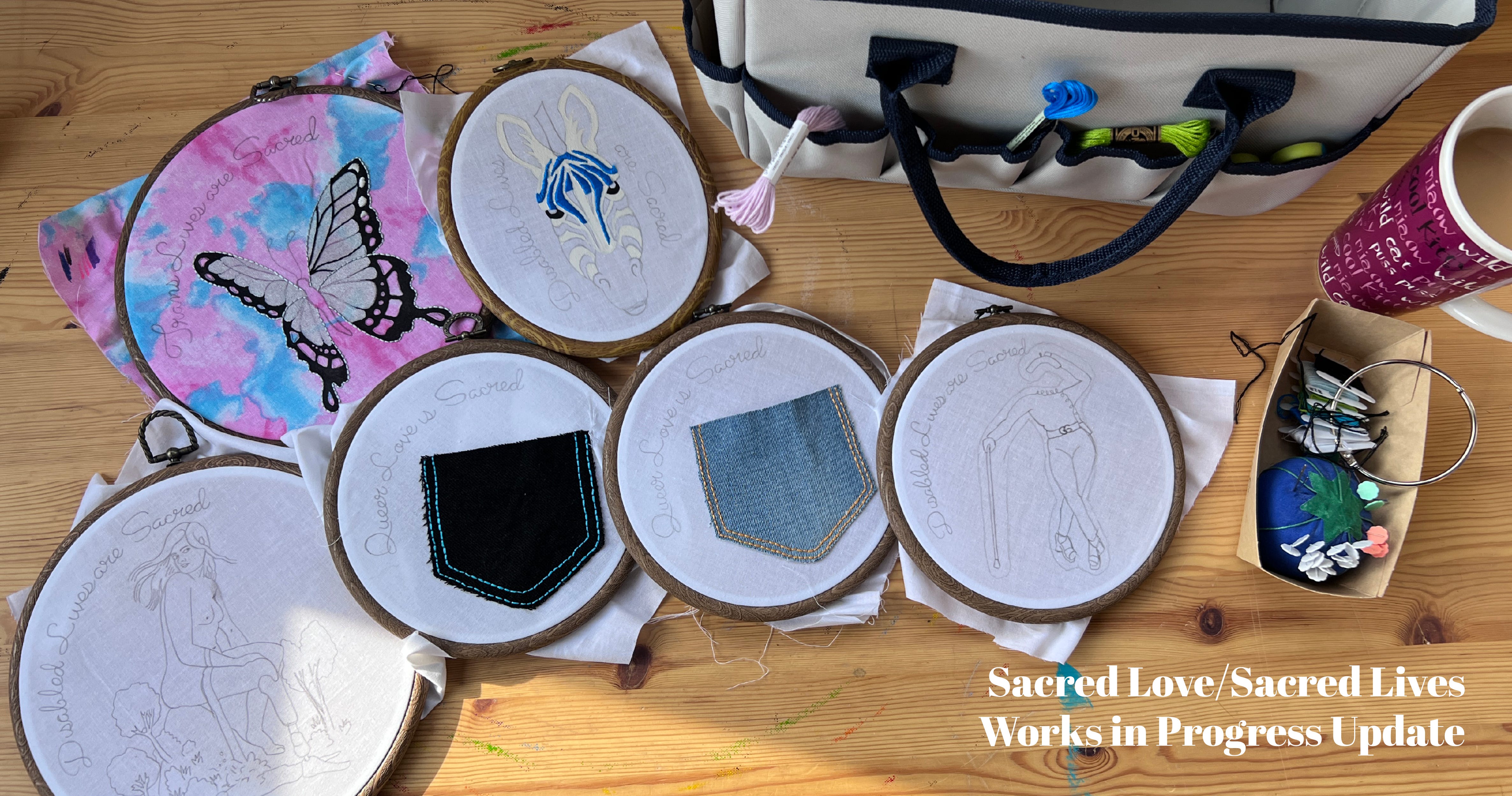 Photo of an array of embroidery hoops on a wood desk top. The hoops are in various shapes and sizes, all with cloth in them and images that are at various degrees of completion. To the right is a mug of tea and a blueberry pin cushion. In the upper right of the image is a grey carry case with many pocket around the outside, embroidery floss hanging from them. 