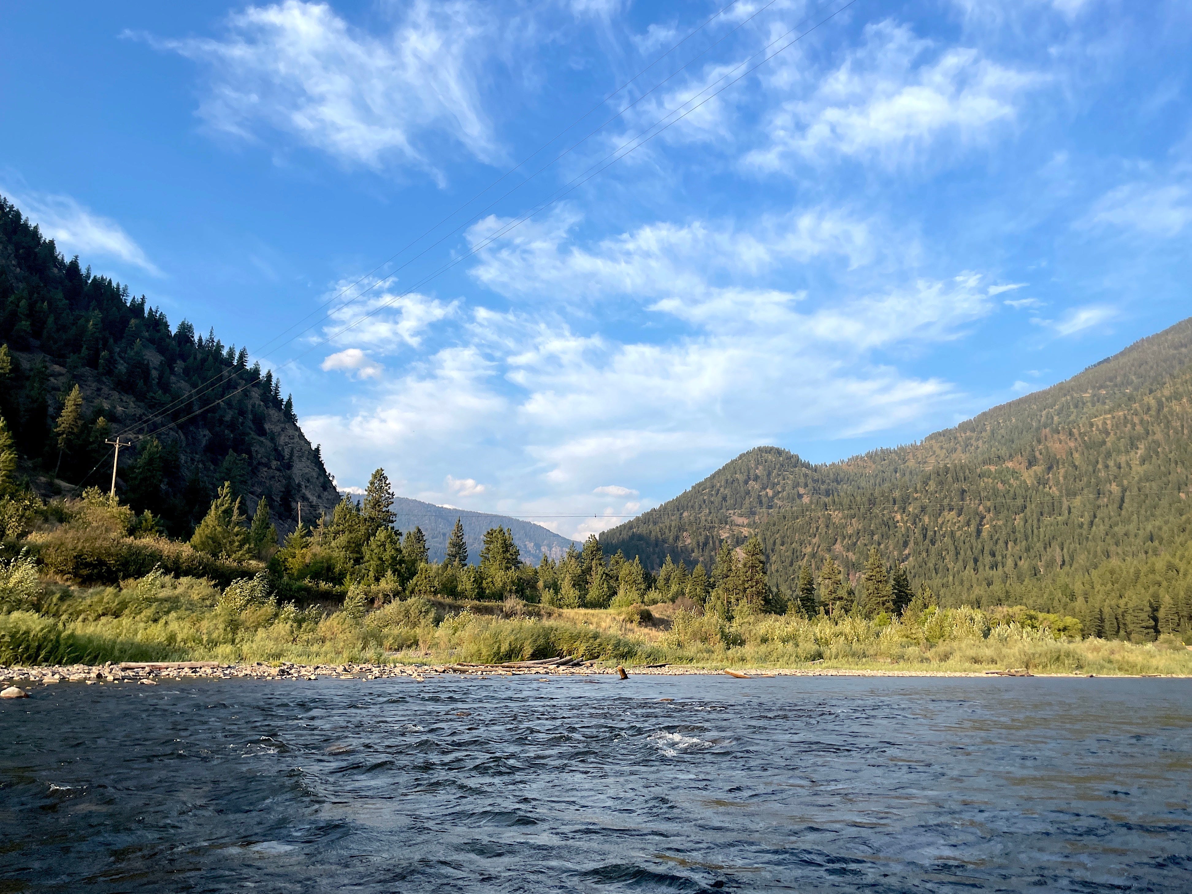A landscape photo of the river behind KettleHouse Amphitheater in Bonner, Montana. A river with mountains on either side dotted with trees, blue sky with wispy clouds, and a power line traversing above the river.