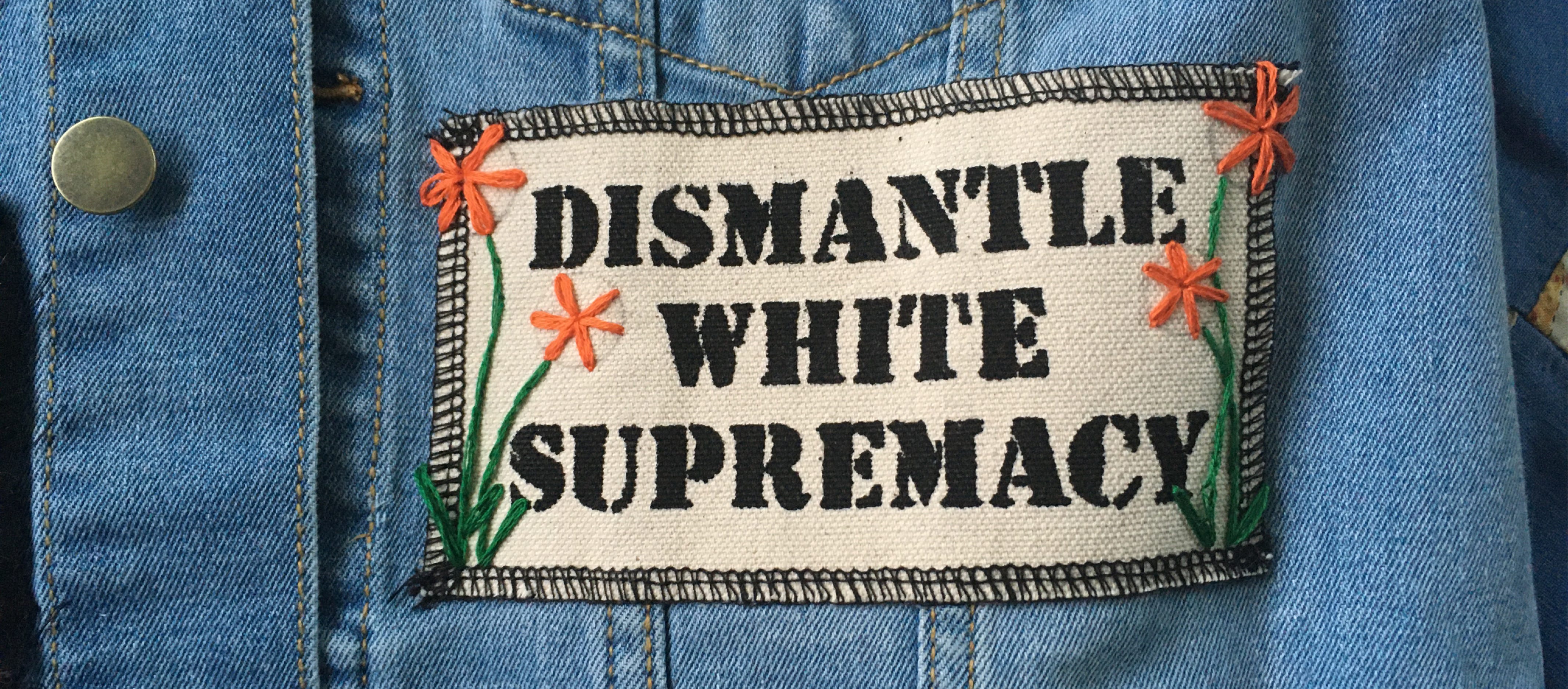 Close up Photo of a white canvas patch sewn onto a light blue denim vest. The patch reads: Dismantle White Supremacy and has been embroidered with little orange flowers along either side. 