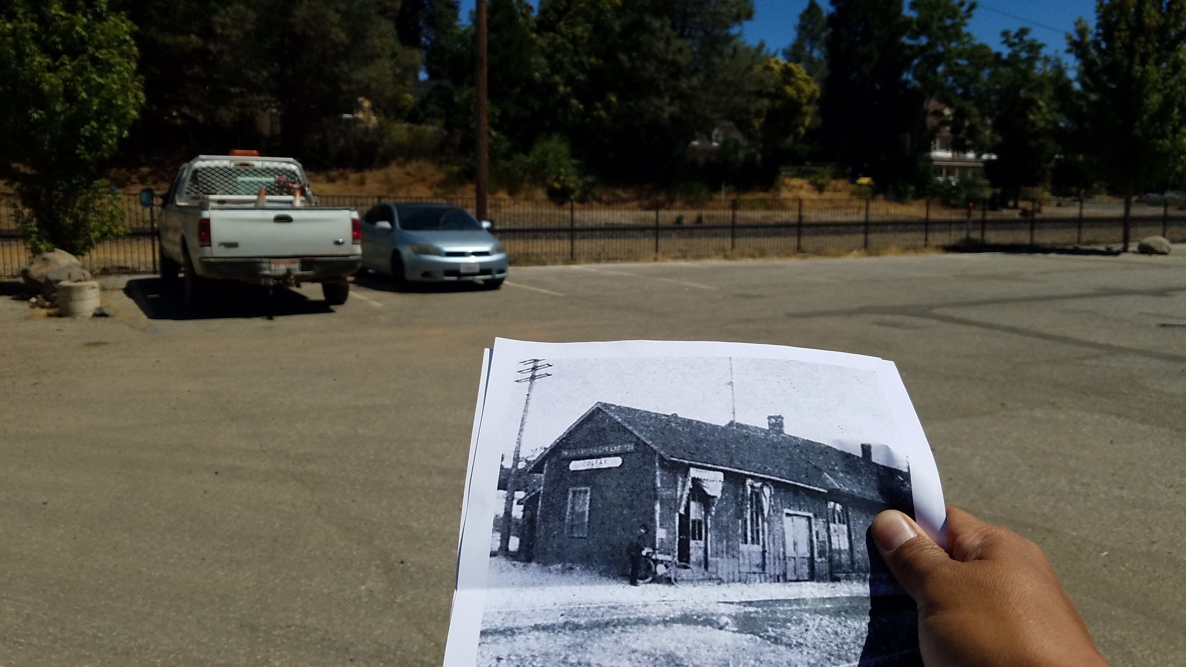 Photo of a parking lot beside railroad tracks. A white pickup and silver sedan are the only two cars present. In the foreground, hand is holding a piece of paper showing an old photo of what the parking lot used to look like. In the photo is an old wooden building with a wooden telephone pole beside it A man has his motor bicycle leaning up against the corner of the building. That is George Wyman.