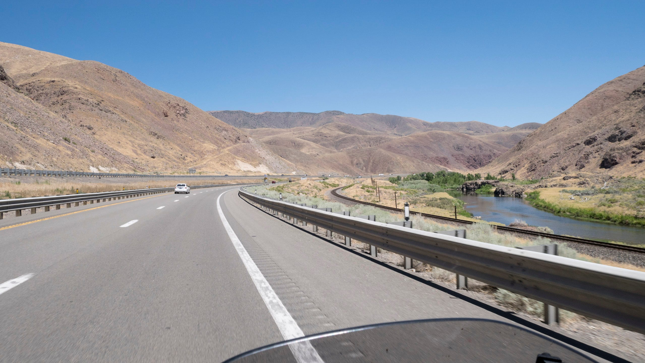 A wide, modern interstate arcs towards distant,  rugged, desert mountains beneath a blue sky. The mountains are brown with very little vegetation. To the right of the highway, beyond the safety guardrail, are a set of railroad tracks also arcing into the distance. To the right of the railroad tracks is a river whose banks are green with vegetation.