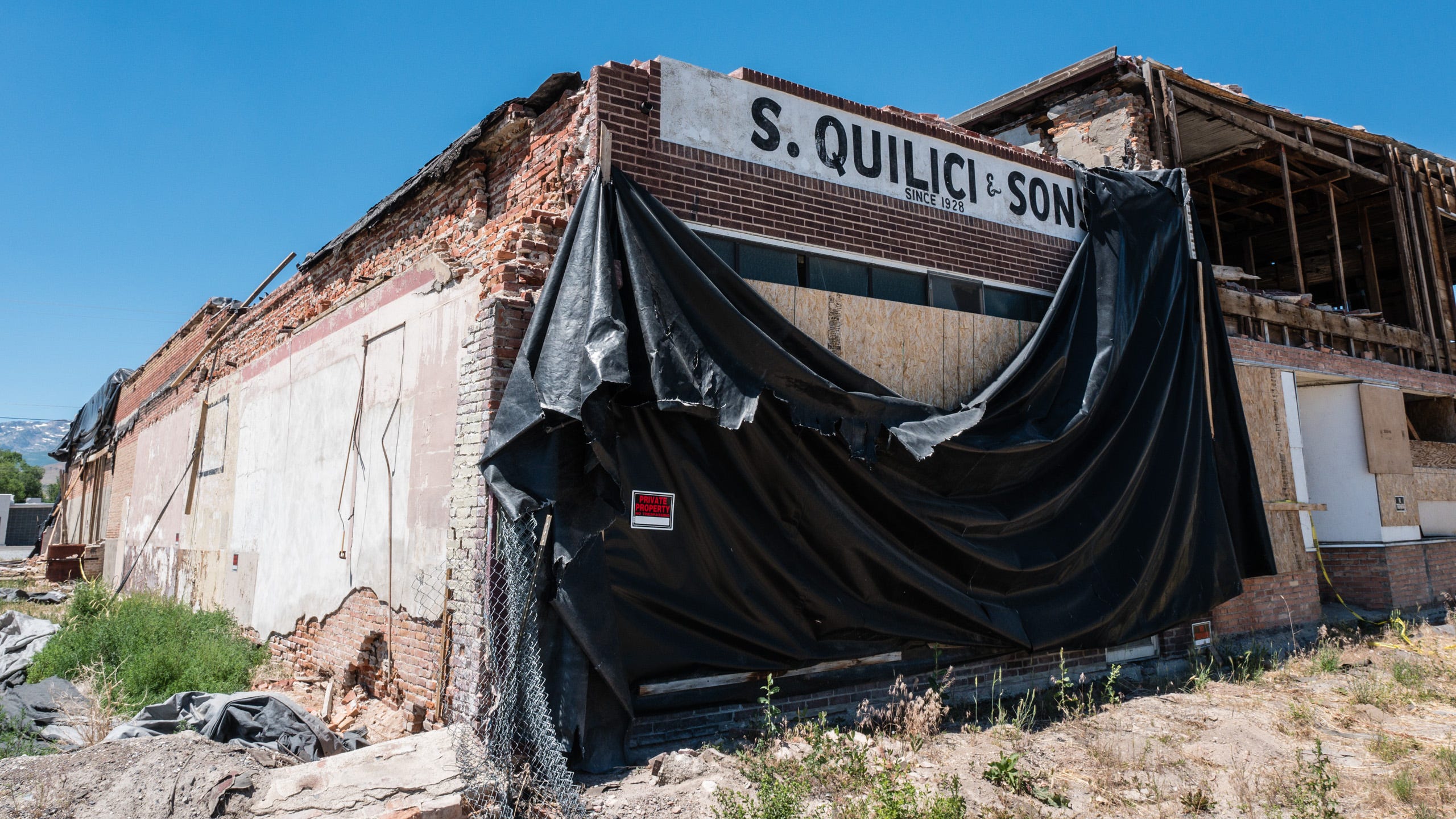 Photo of a red brick building damaged by the 2008 earthquake. The front of the building is covered with a large black tarpaulin that is sagging in the middle, revealing plywood covering the front windows. Above, "S. Quilici & Sons, Since 1928" is painted in black letters on white masonry. The top of the building is shorn off. To the left, the now-exposed side of the building reveals crumbling bricks along the top and bottom of the building, and what was once an adjacent building is now a field filled with debris and wild grasses. A "Private Property" sign is fastened to the large black tarpaulin. To the right is another destroyed building, two stories with plywood covering windows on the first floor and the second story facade completely gone, revealing the studs and joists of the building.