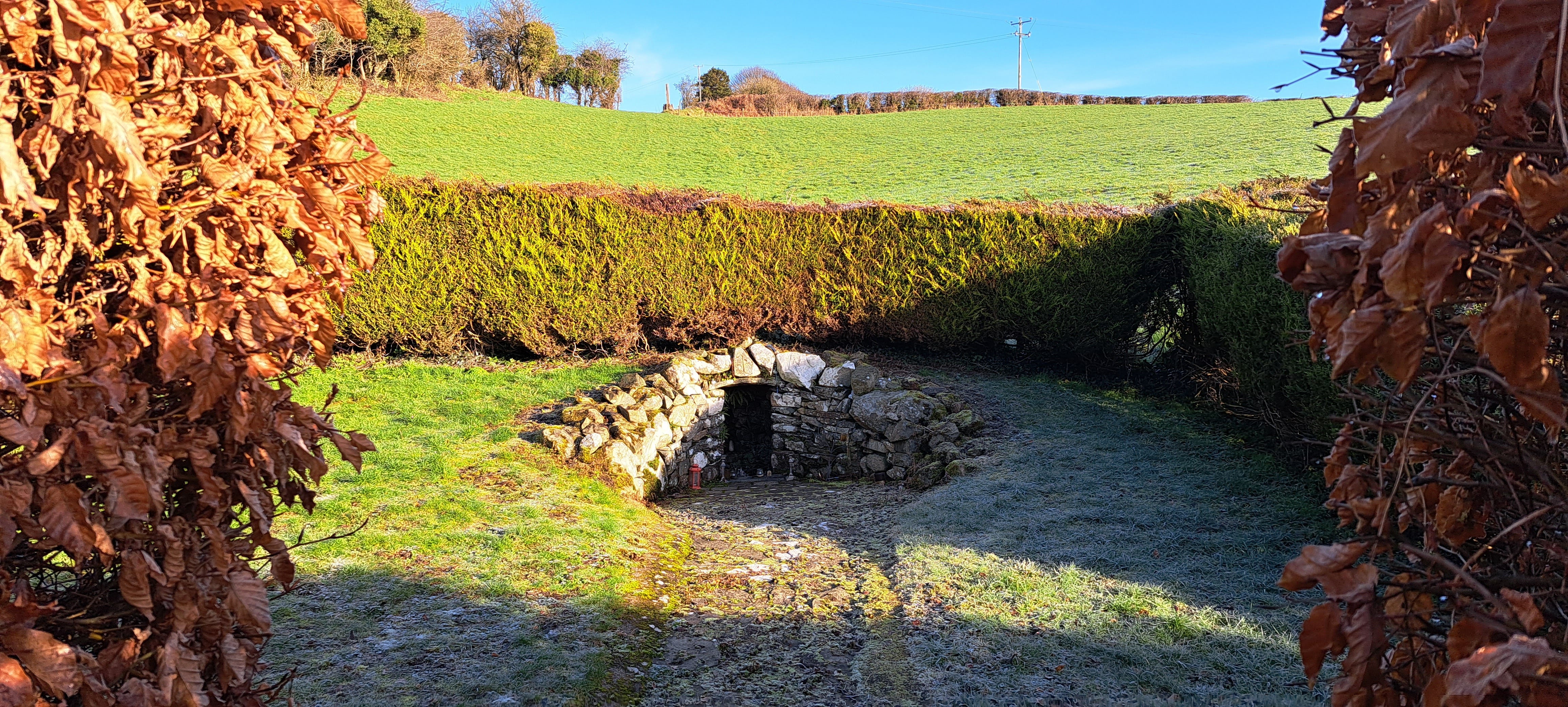 Looking through a gap in the hedge at a holy well set below a hill; the well is built into the hill with a rough stone facade arched with an opening to the water, its almost like a tiny cave in the hillside.