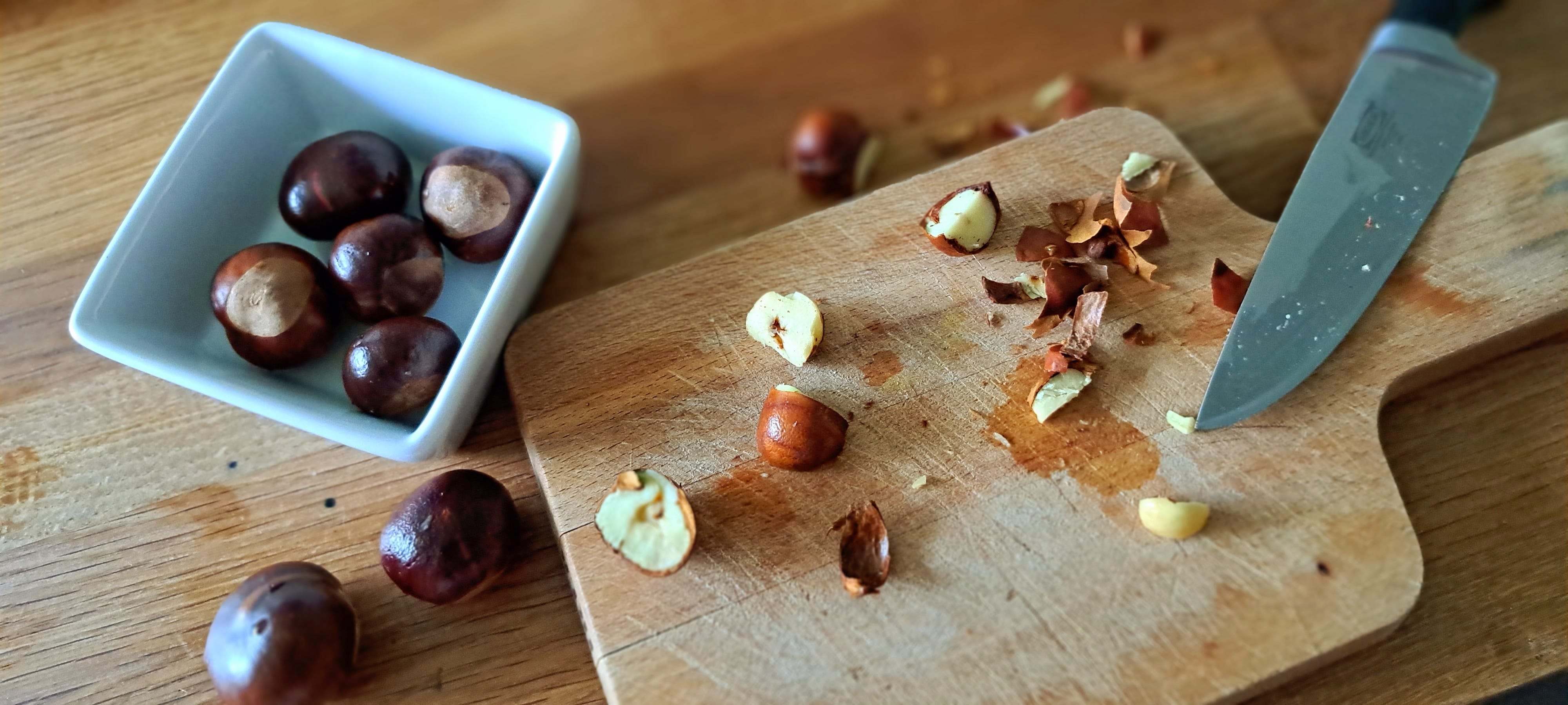 Chopped up conkers on a chopping board, with a knife to the right, a square white ramekin on the left containing whole conkers, shells and papery brown skins scattered on the wooden worktop.