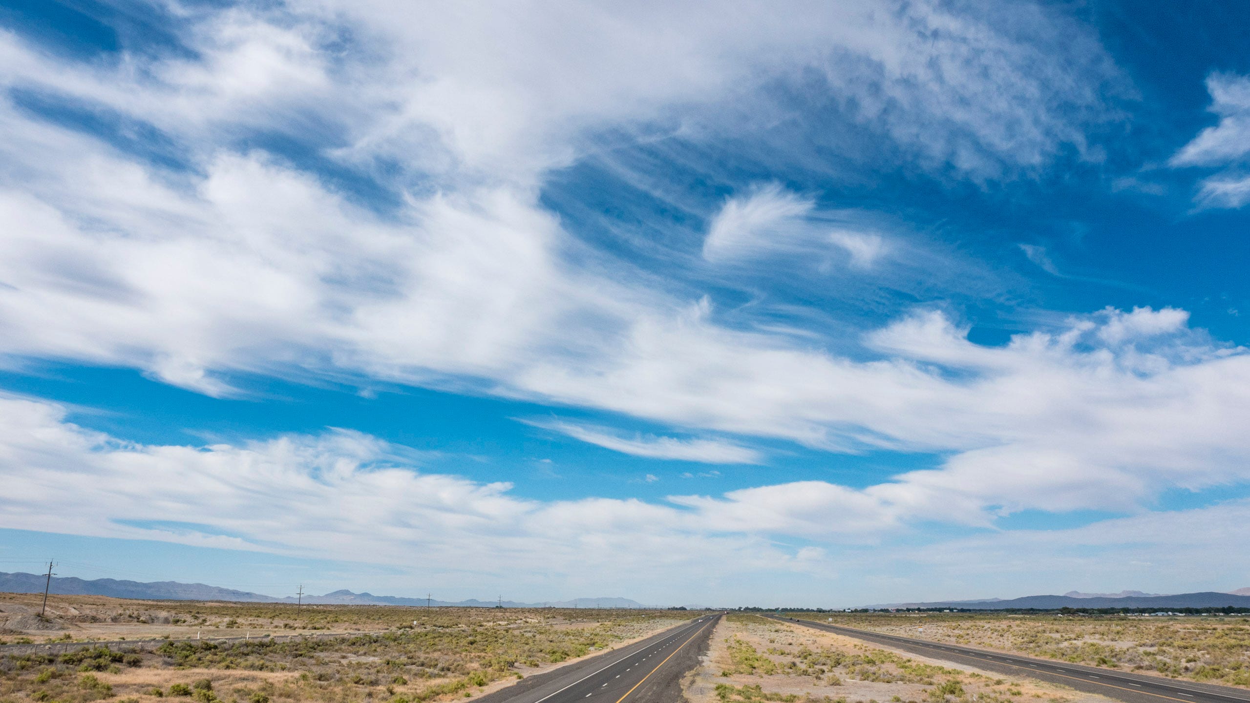 Photo of clouds floating in a blue sky over the flat desert of Nevada. They sky is a cool, blue, the clouds white and whispy. The sky takes up the upper 80% of the photo while the expansive desert of Nevada occupies the lower 20%. The desert is dotted with faded green brush. The interstate cuts across the desert to a vanishing point on the distant horizon, where the silhouettes of jagget mountains can be seen. The land is dwarfed by the sky.
