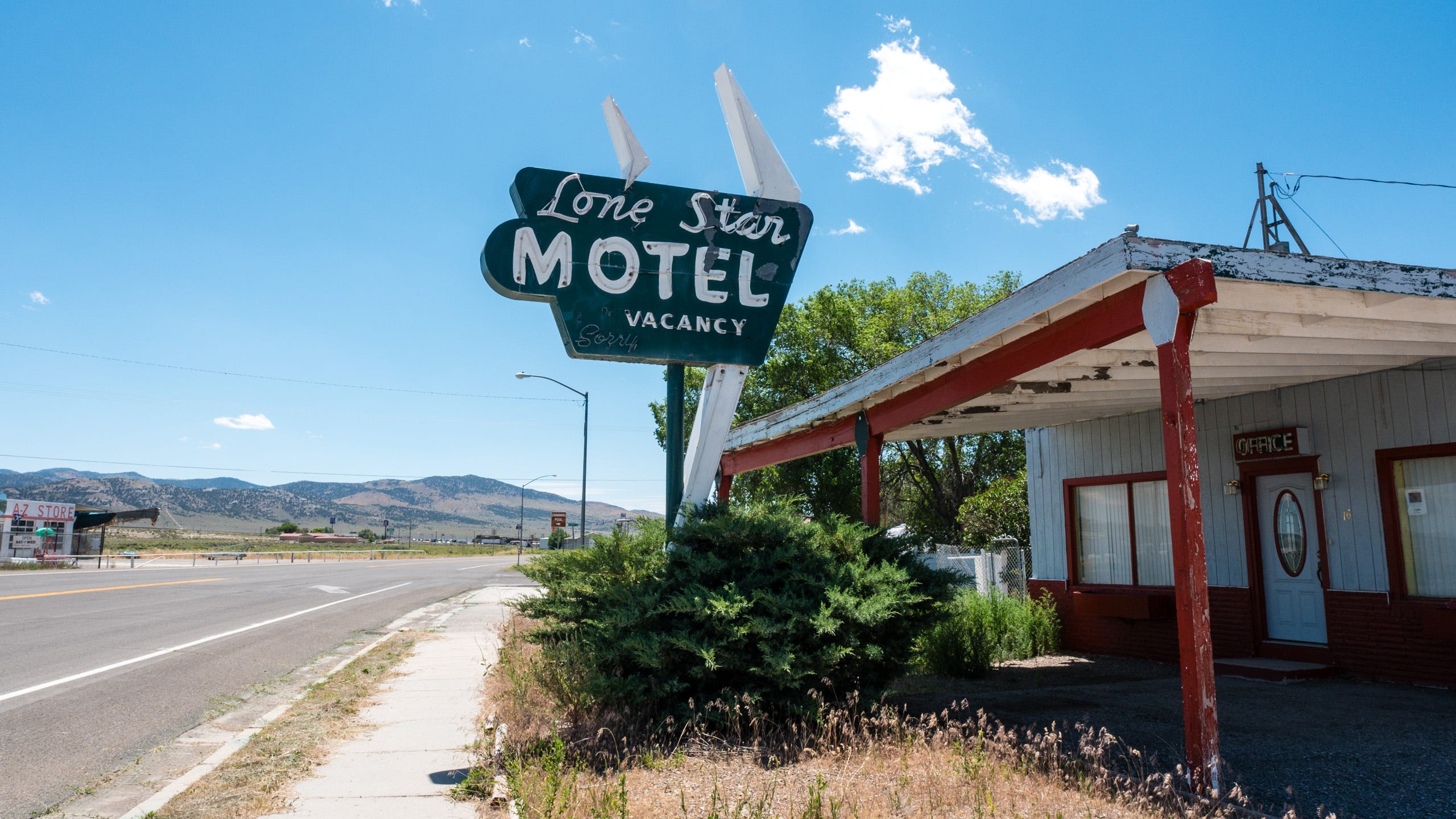 Photo of the Lone Star Motel, an abandoned mid-century roadside motel. The "Lone Star Motel" sign is mid-century futuristic, like something from the Jetsons, with angular lines and rounded corners. "Lone Star" is in white script and "Motel" looks like it was written with a white magic marker. The neon tubes are visible but unlit and they are set in a faded green field. Below is "VACANCY" with space to the left to add "NO" and script letters below that say, "Sorry,". To the right, is the covered patio, the sagging white roof held up by simple red wooden posts whose paint is peeling. The patio leads to the OFFICE, another neon sign, white letters upon a red field. The shrub and grasses at the base of the sign are unkempt. To the left, the road leads to mountains in the distance.