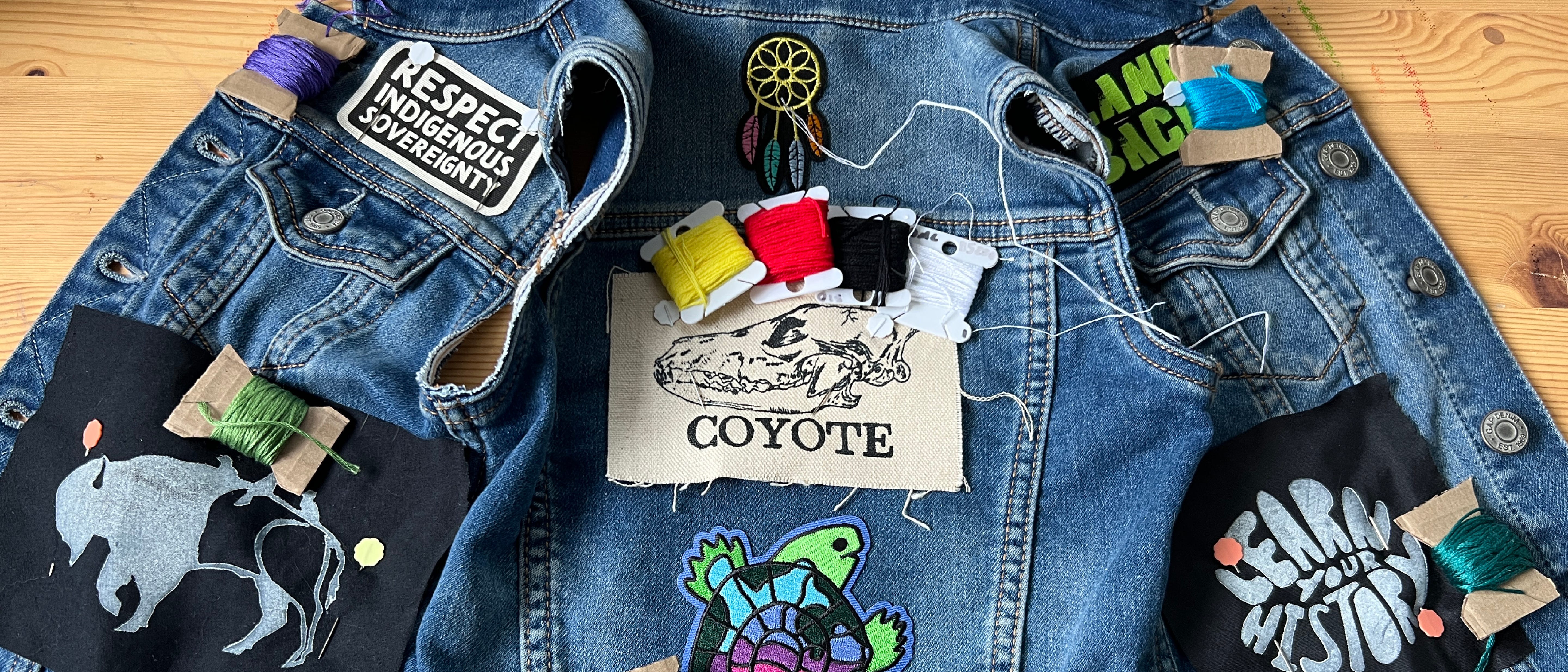 Photo of a denim vest spread out on a wooden desk with patches laid out on it next to embroidery floss on bobbins to be used on each patch. The patches follow an Indigenous resistance theme, withe slogans like "LandBack" and "Respect Indigenous Sovereignty". 