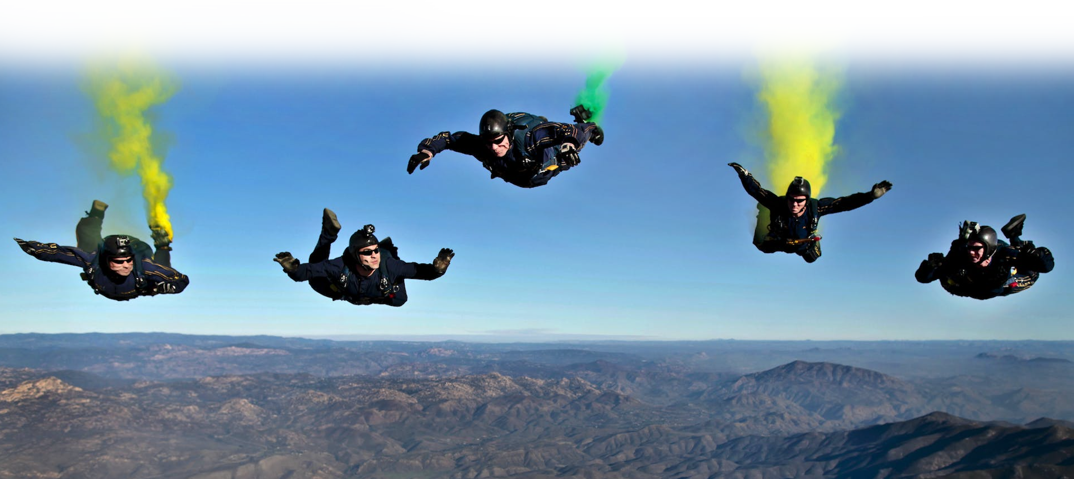 A group of sky divers with green and yellow smoke coming out of their unopened parachute packs.