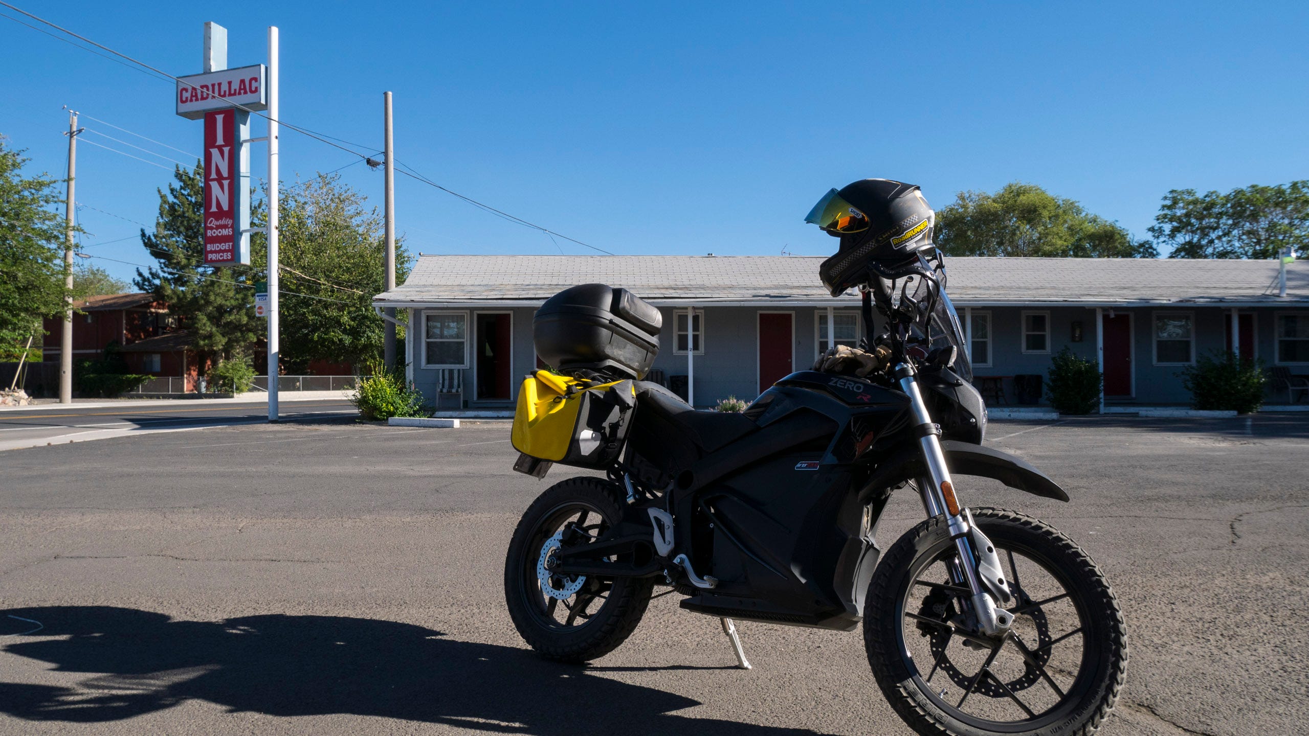 Photo of a black electric motorcycle with yellow bags in the parking courtyard of the Cadillac Inn. It's a sunny day but the sun is lower in the sky, causing the motorcycle to cast a long shadow on the asphalt. On the far side of the courtyard is one of the low-slung, one-story wings of the motel, with a nearly flat roof and red doors to the rooms opening up to the courtyard. By the street is the Cadillac Inn sign on a tall white pole.