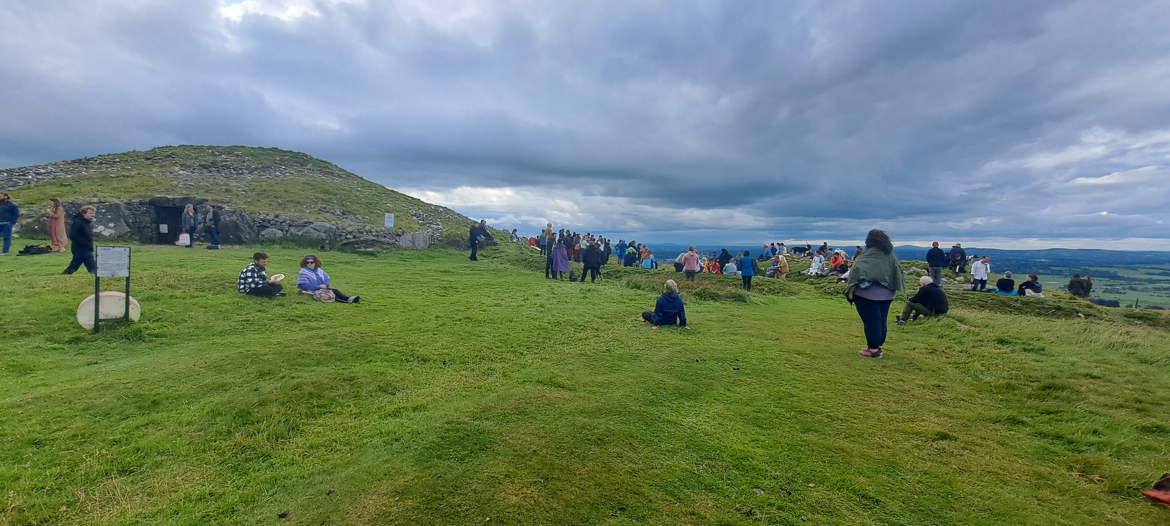 People beginning to gather at Loughcrew; the mound of Cairn T is to the left of the image beneath a dark, cloudy sky against which the green of the grass seems very bright. In the foreground and to the right of the image, groups of people begin to gather and wait.