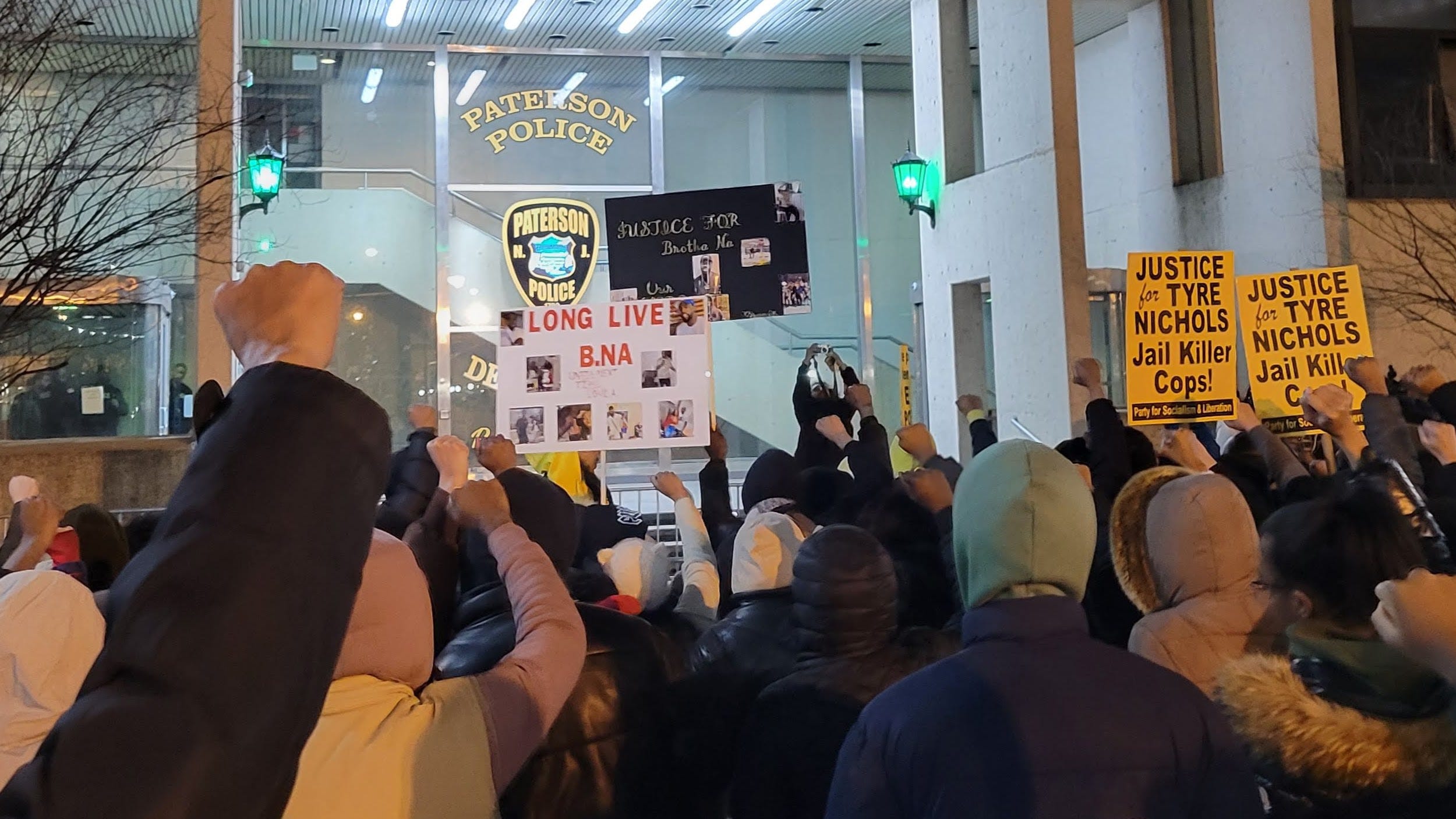 Protesters hold signs supporting Najee Seabrooks and Tyre Nichols while holding their fists in the air in front of the Paterson Police Department
