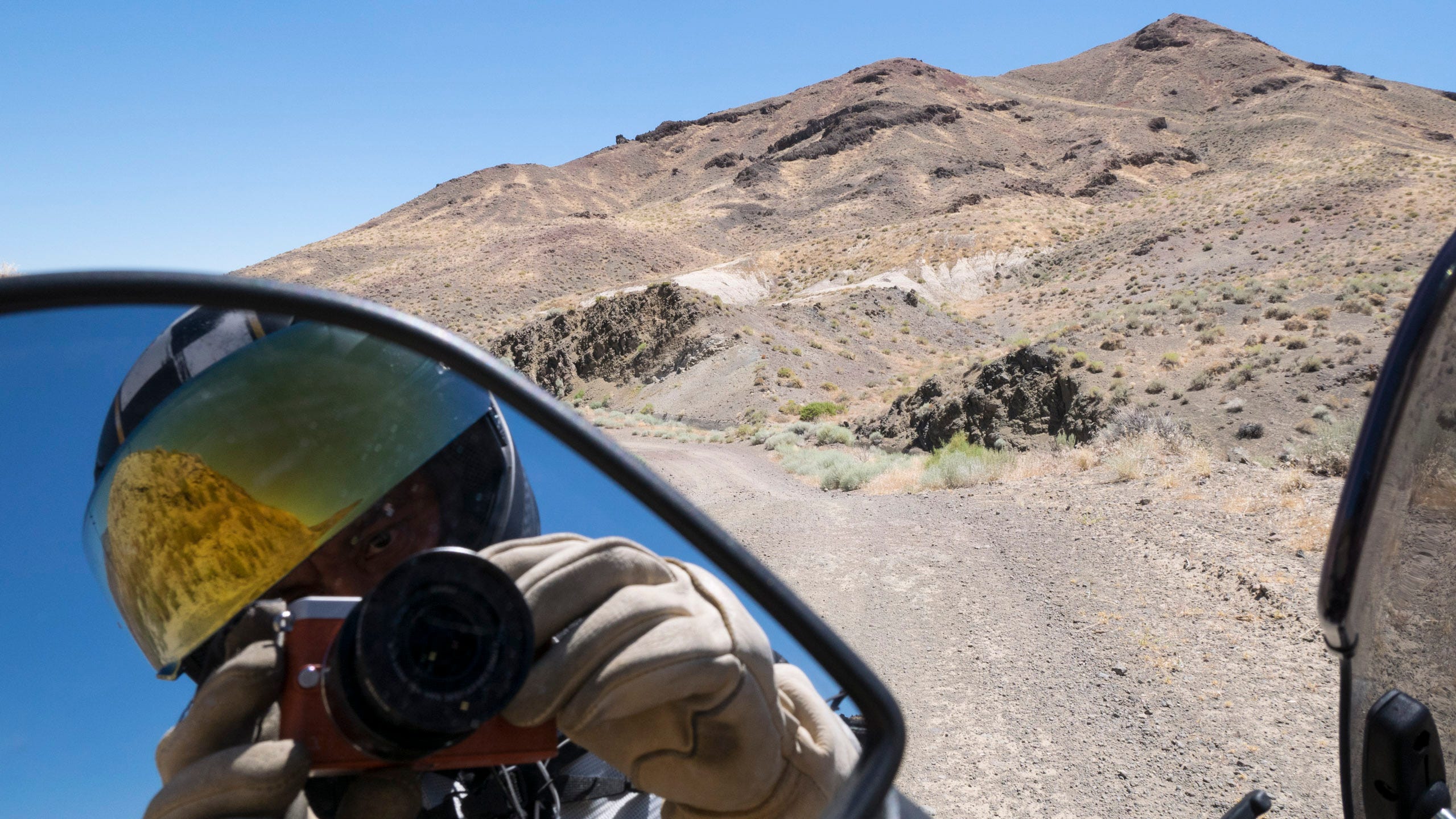 Photo from the seat of a motorcycle, including the land ahead of the motorcycle, a piece of the windshield on the right edge of the photo, and the author reflected in the left rearview mirror in the lower left corner of the photo. The land ahead of the motorcycle is a tall, naked mountain in the desert with multiple, weather-worn peaks leading up to a naked top. Small clumps of grass grow in patches and exposed, dark rock appear on the hill in places. The dirt road that the motorcycle on bend to the left. The author reflected in the mirror of the motorcycle is wearing elkskin leather gloves and a black helmet with a checkerboard strip pattern along the top and a gold reflective visor that has been flipped up a little for ventilation. The reflection in the visor is more scrub brush growing in the sandy soil and the blue sky, both tinted gold. The author is holding a small orange camera with a black lens that is being used to take the photo.