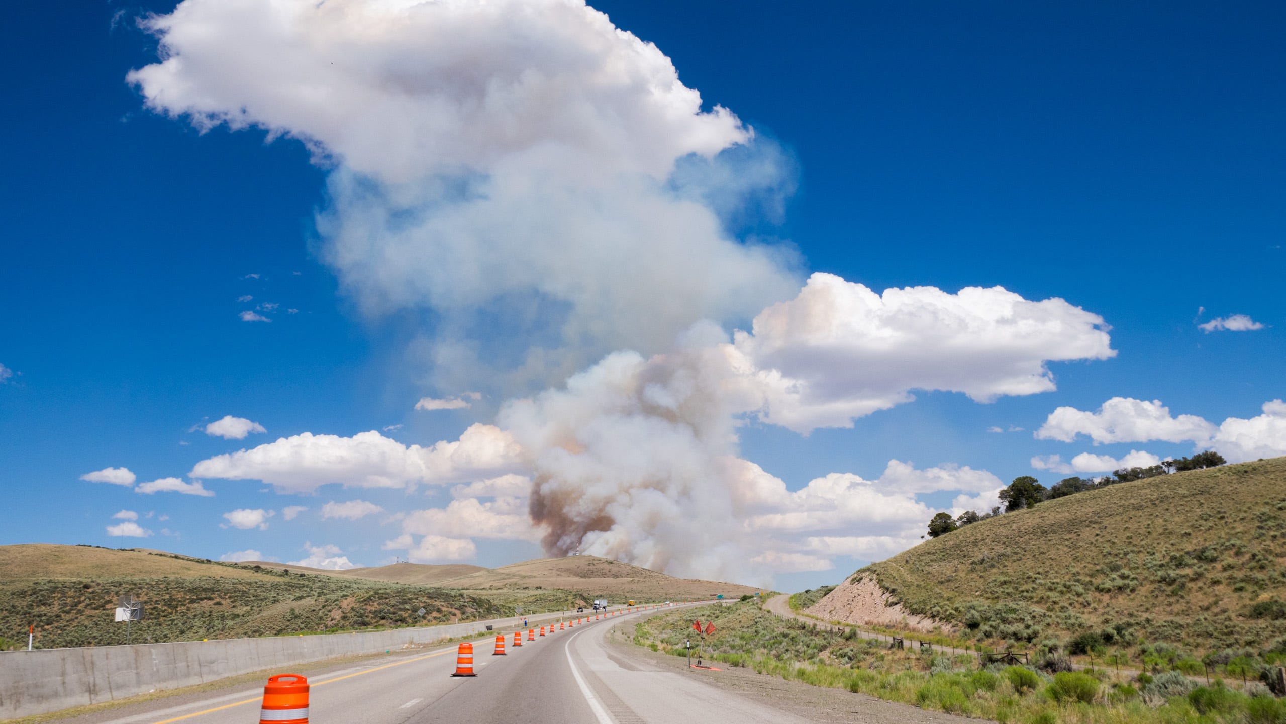 Color photo of a road climbing a grade to get over a mountain. The mountain is mostly treeless but there are dry brown grasses. The road bends to the right as it climbs. To the left, behind a rise, brown smoke from a forest fire rises to the sky to meet big white clouds.