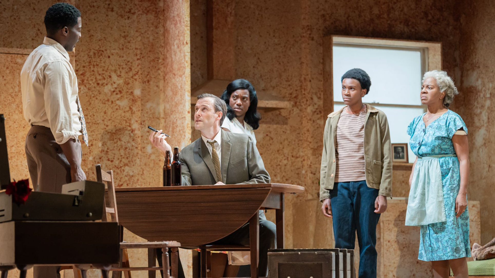 A white man in a rumpled business suit sits at a bare kitchen table offering a pen for signature to a black man who is surrounded by his horrified-looking family.