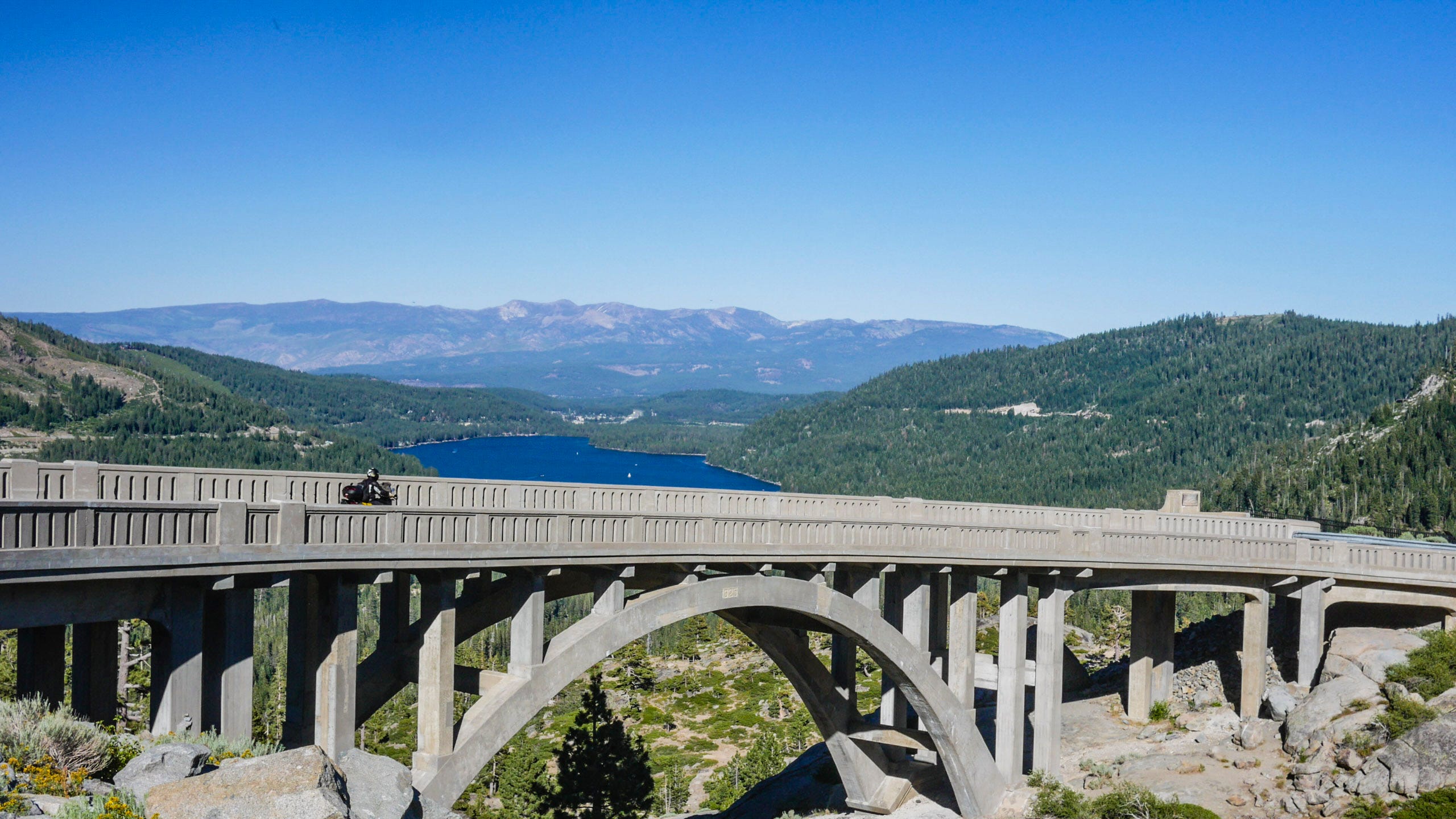 An older arched concrete bridge crosses a rocky chasm from the left and descending to the right. A lone motorcyclist descends on the bridge. Beyond, the deep blue waters of Donner Lake are framed by rolling green mountains. And in the hazy distance lie more purple mountains. On the mountain on the right, you can see a cut along the mountain that is the Transcontinental Railroad.