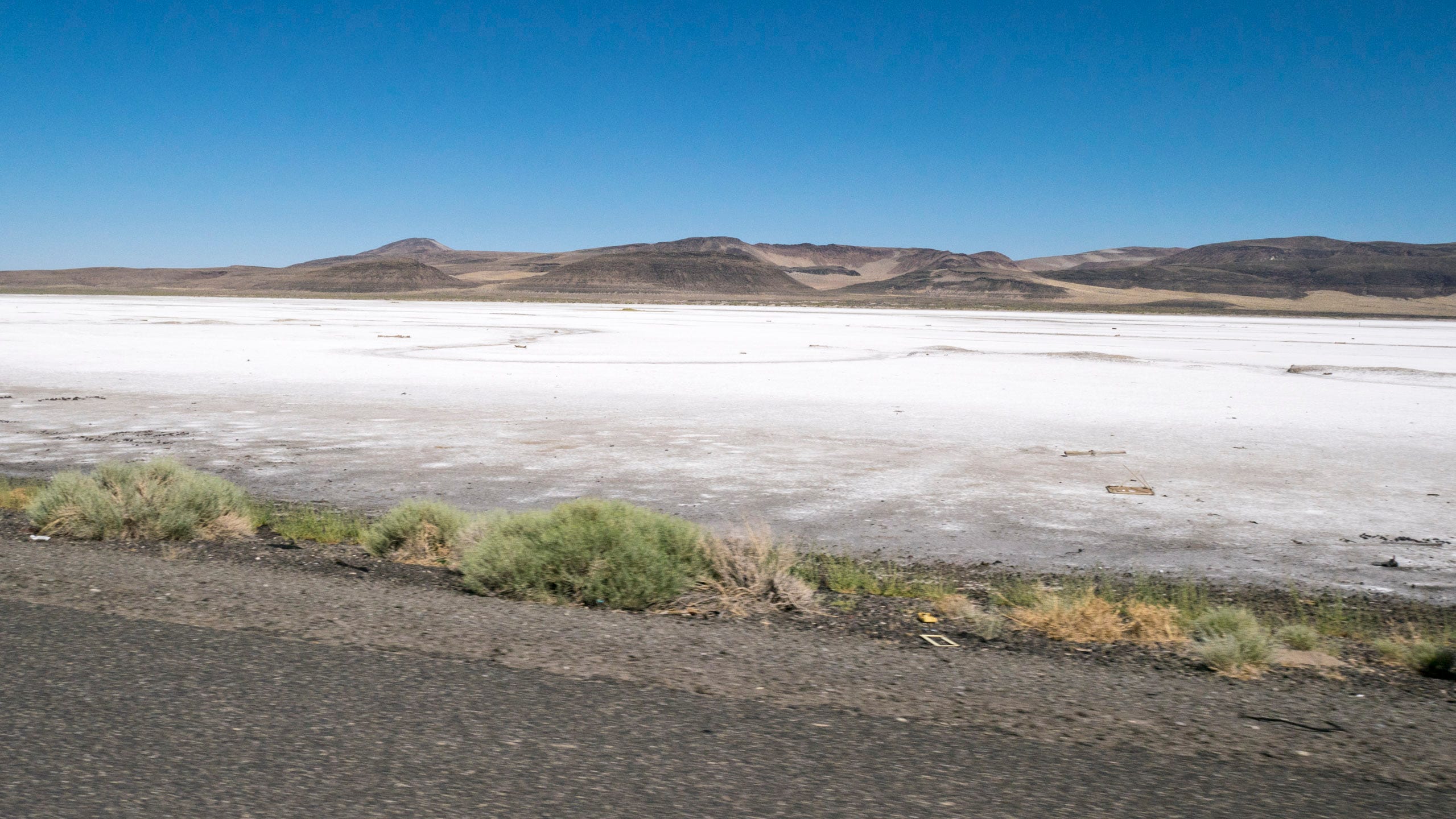 Photo of a white, sandy desert running along the side of the highway. The desert fills the width of the photo and appears to be a mile or more wide. Naked, rolling mountains lie on the horizon.
