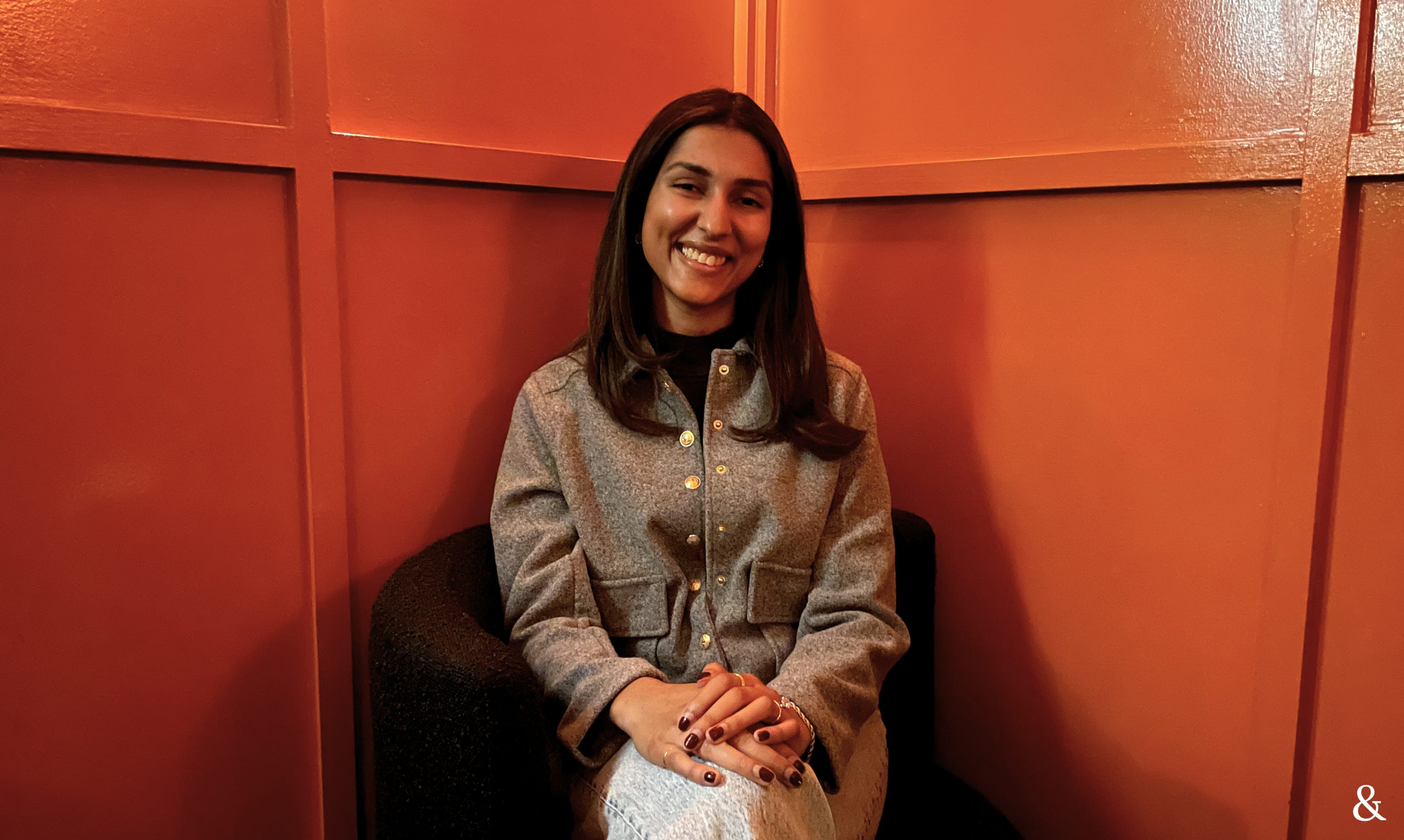 Abi Suresh in a gray jacket sitting and posing facing the camera in front of an orange wall.